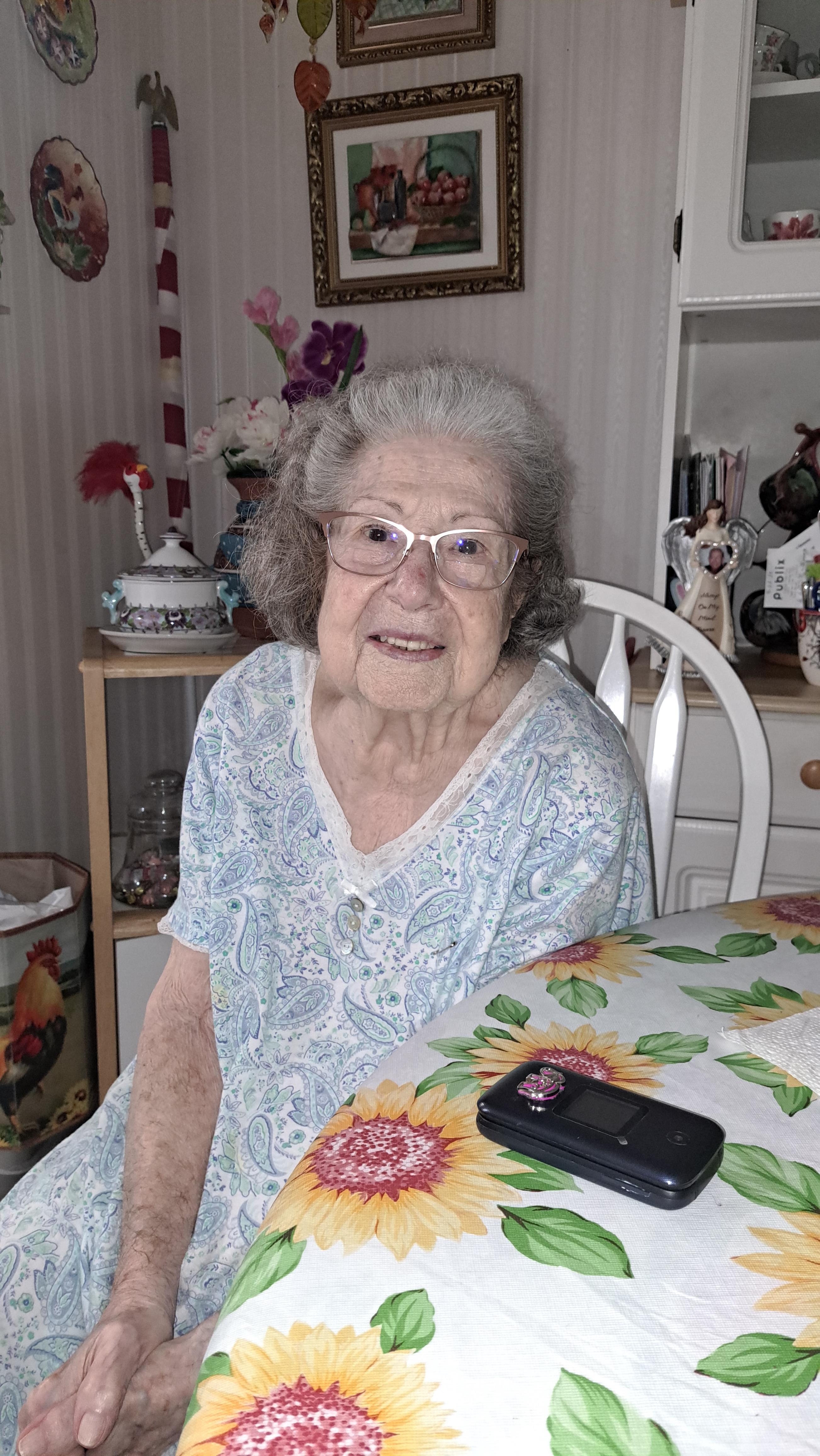 An elderly woman sits comfortably at a table adorned with flowers, radiating joy and warmth.