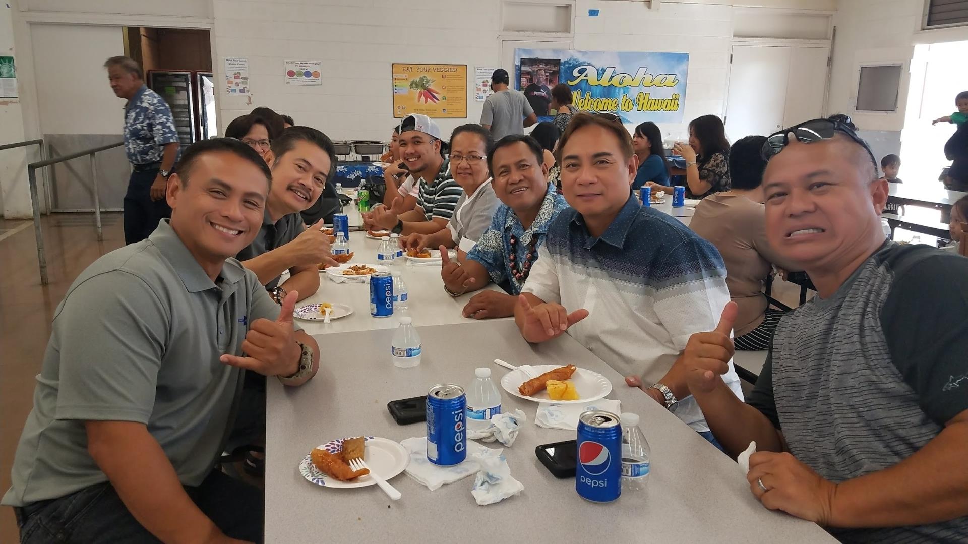 Friends laugh and share food while sitting at a long table in a lively community space.
