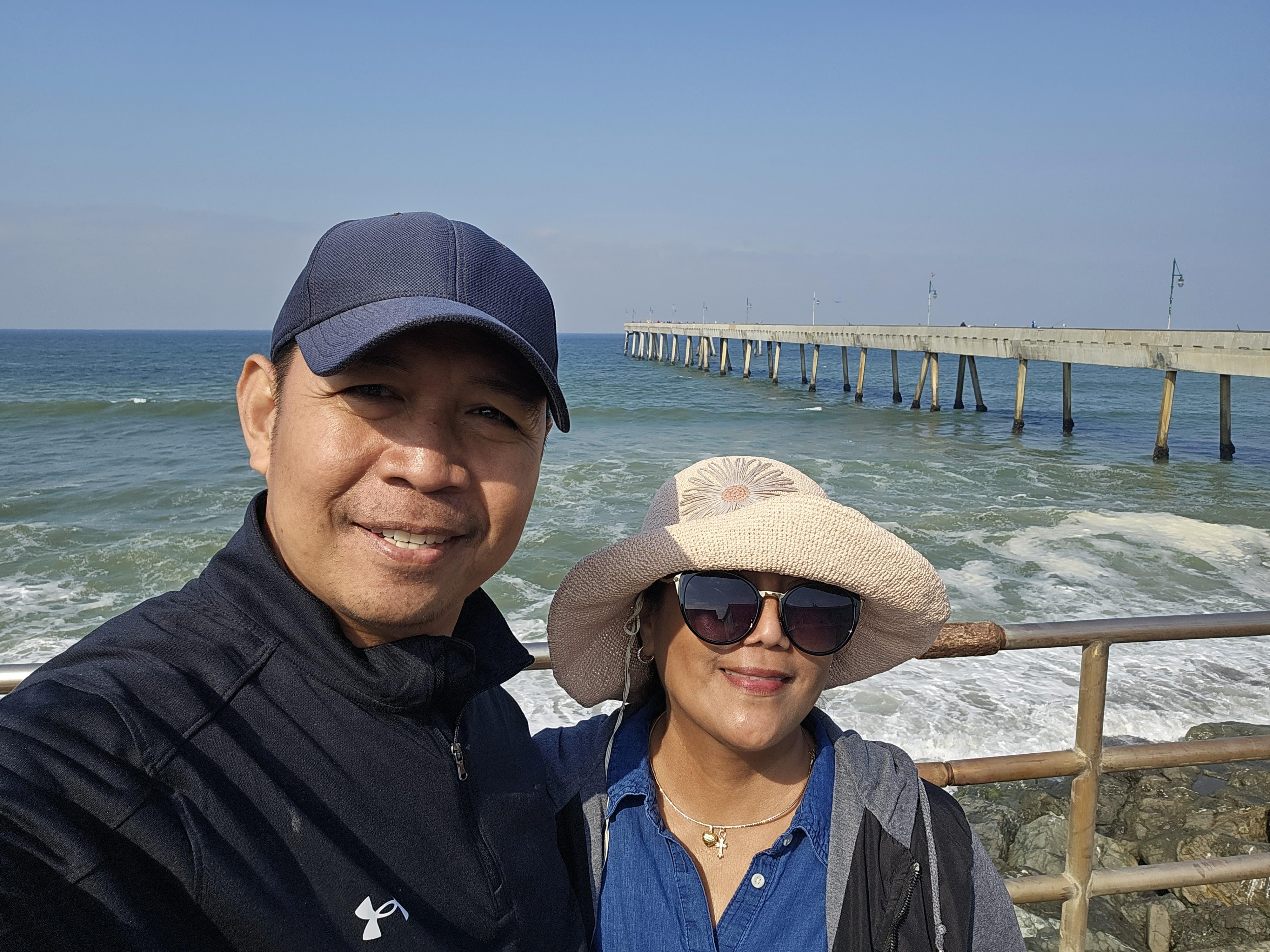 A couple stands together smiling at a coastal pier under bright blue skies and ocean waves.