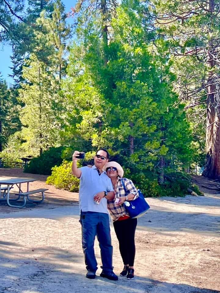 A man and woman smile together while capturing a selfie in a sunny forest with tall trees.