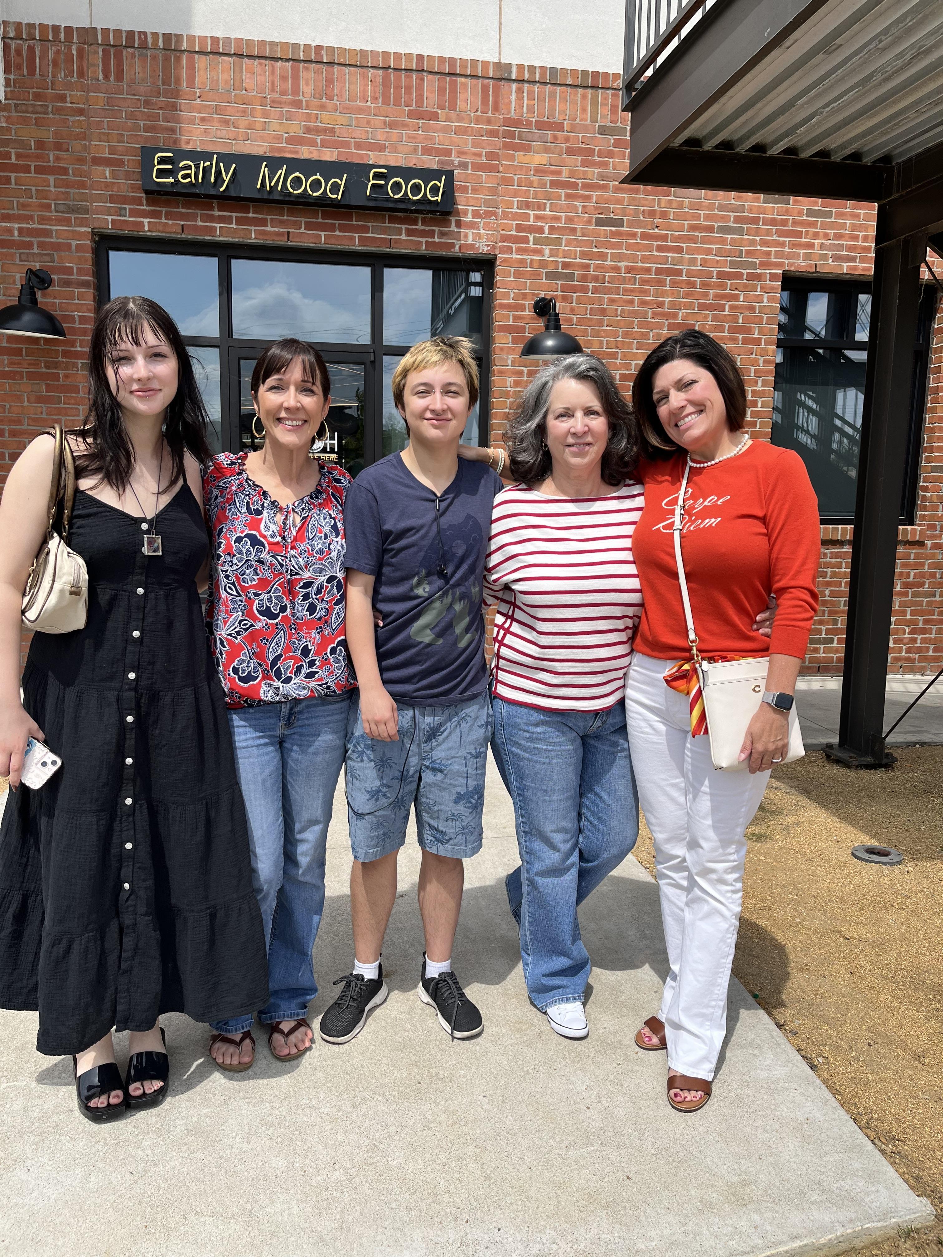 Friends gather in front of a restaurant, smiling and enjoying a sunny day together.