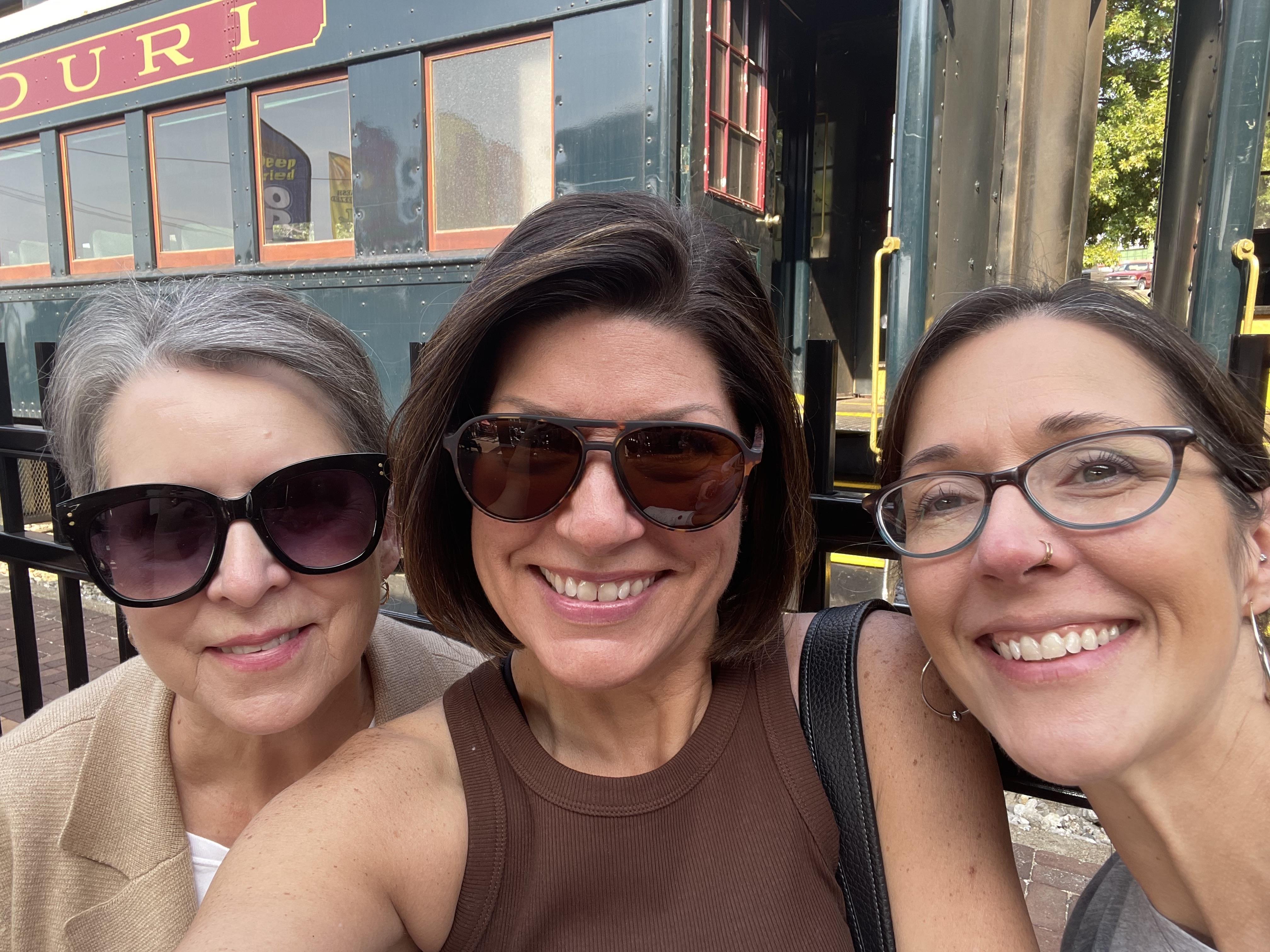 Three women are smiling widely, posing together outside a vintage train on a sunny day.