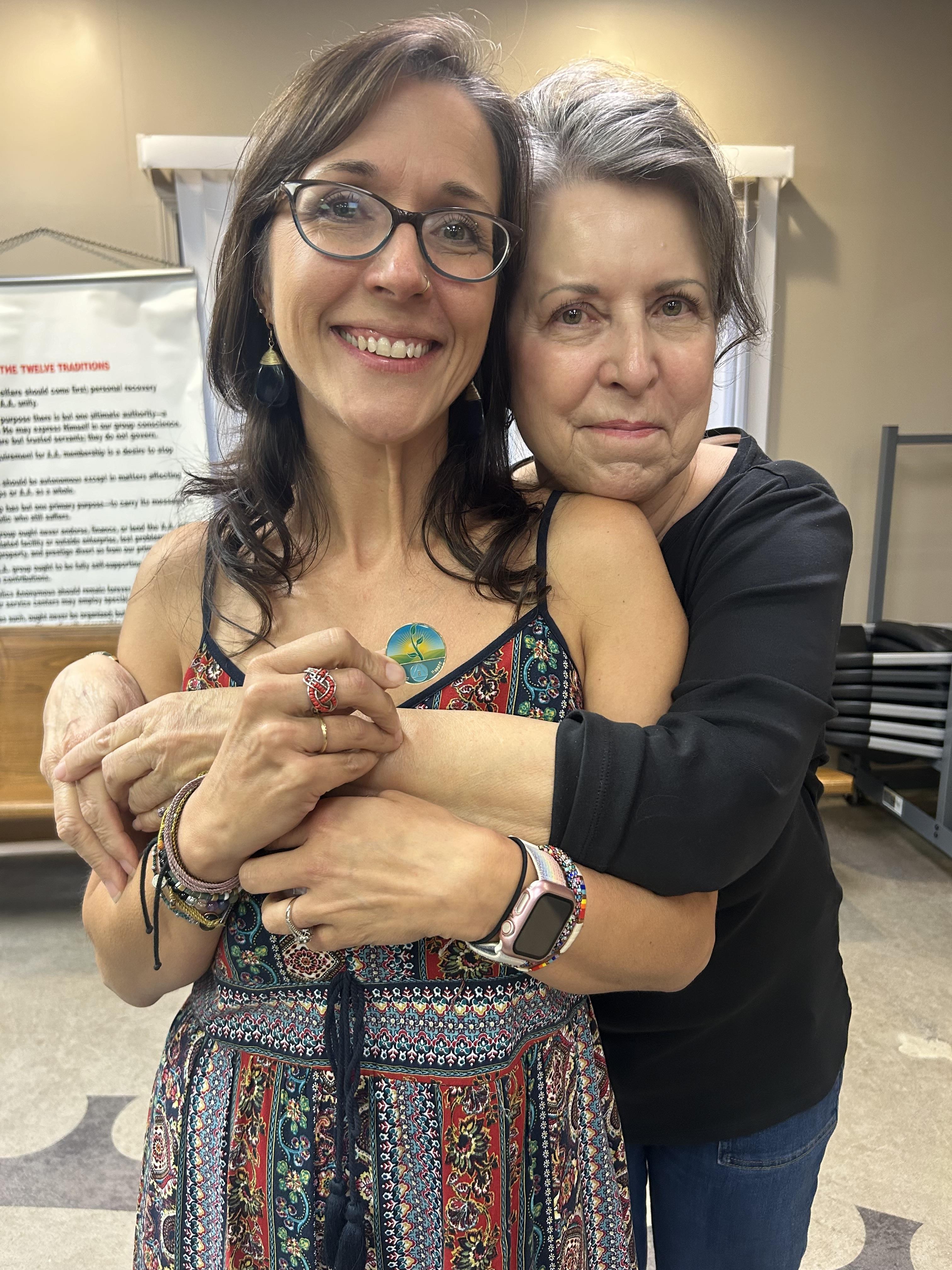Two women share a warm embrace at a community gathering, smiling and enjoying the moment together.