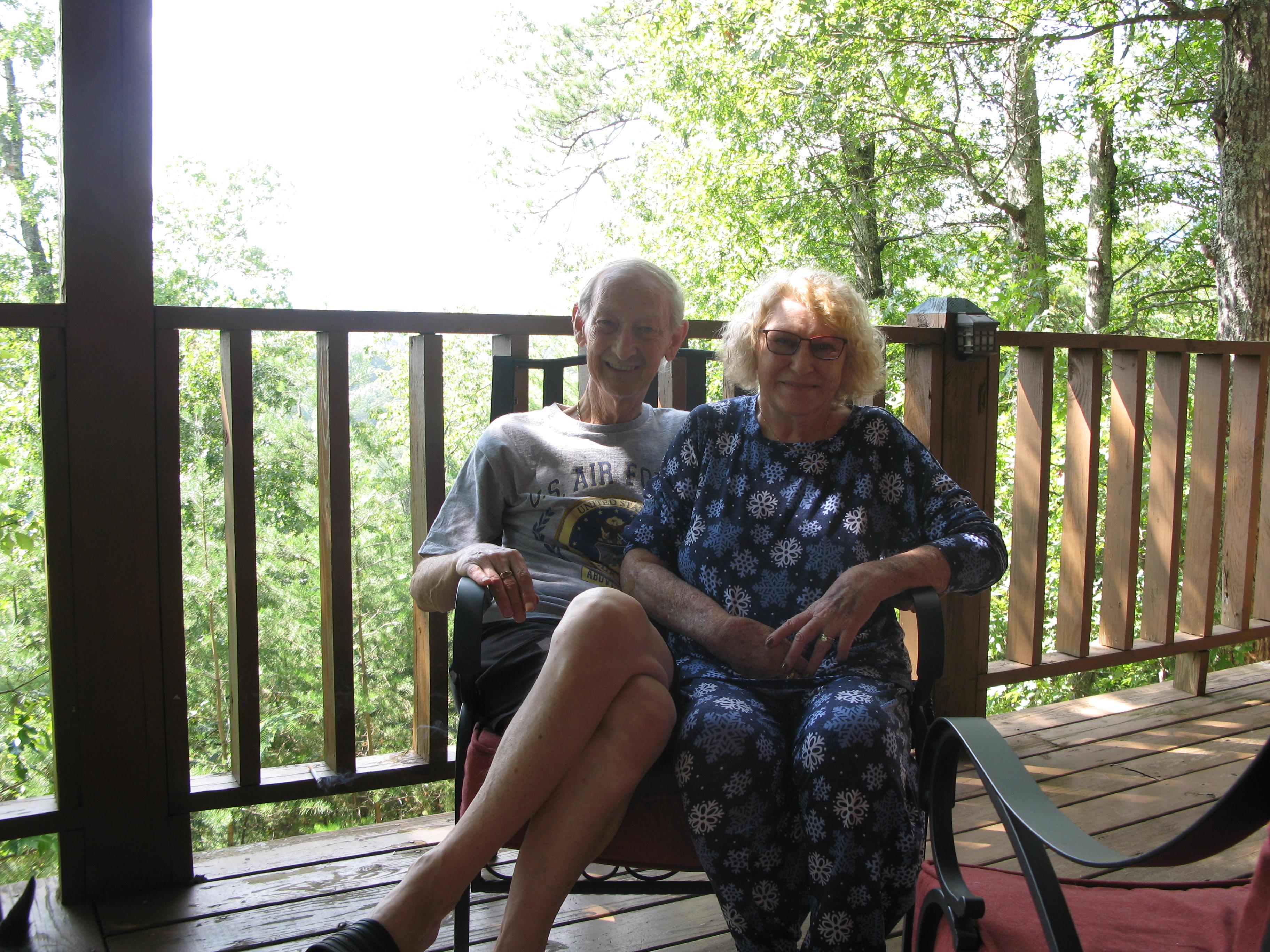 Elderly couple relaxes together on a wooden deck, enjoying a sunny summer day surrounded by nature.