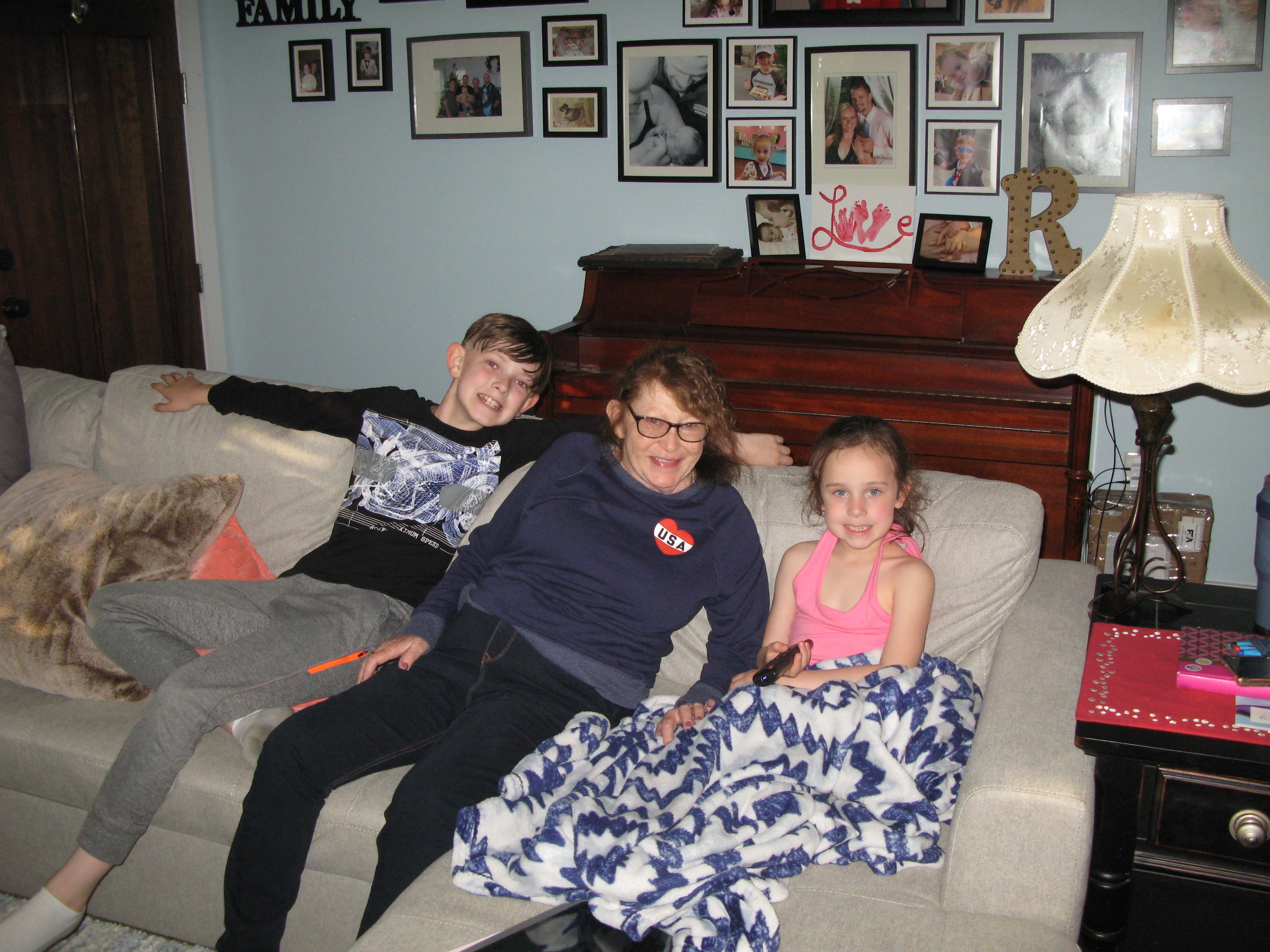 Three children relax on a couch with blankets in a cozy living room adorned with family pictures.