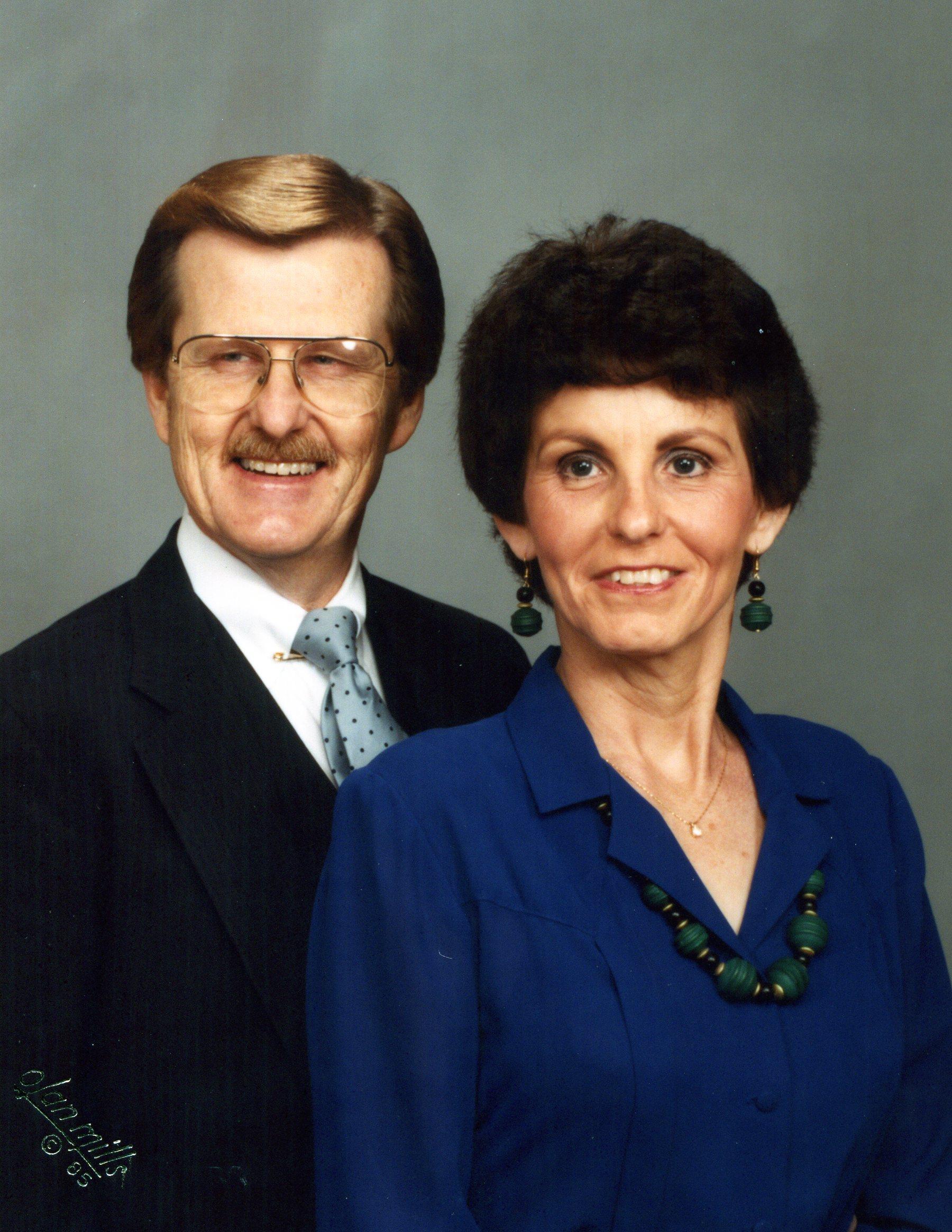 A couple stands together in a studio, both dressed formally and smiling warmly at the camera.