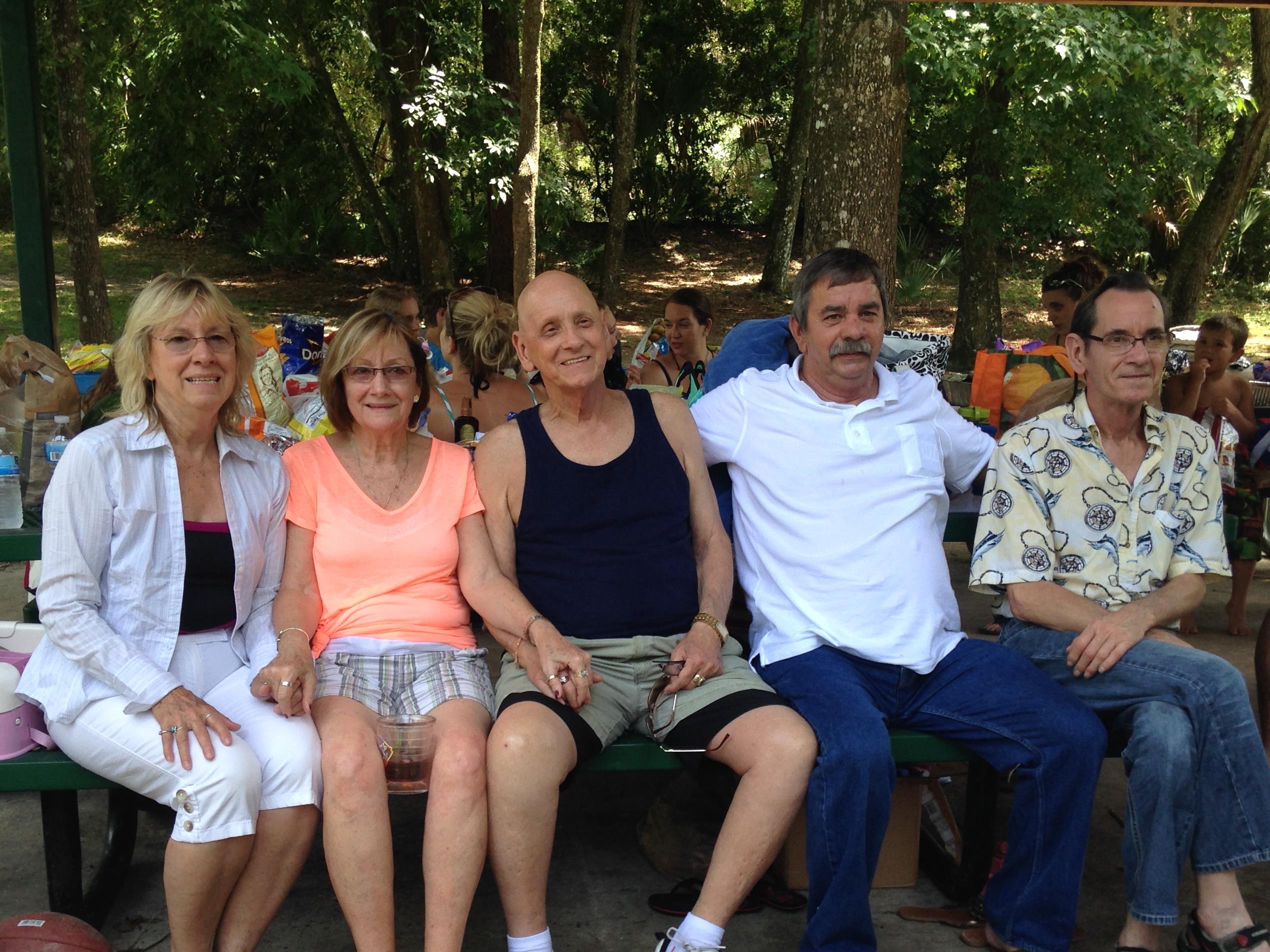 Five friends sit on a bench outdoors, smiling and enjoying each other's company under the trees.