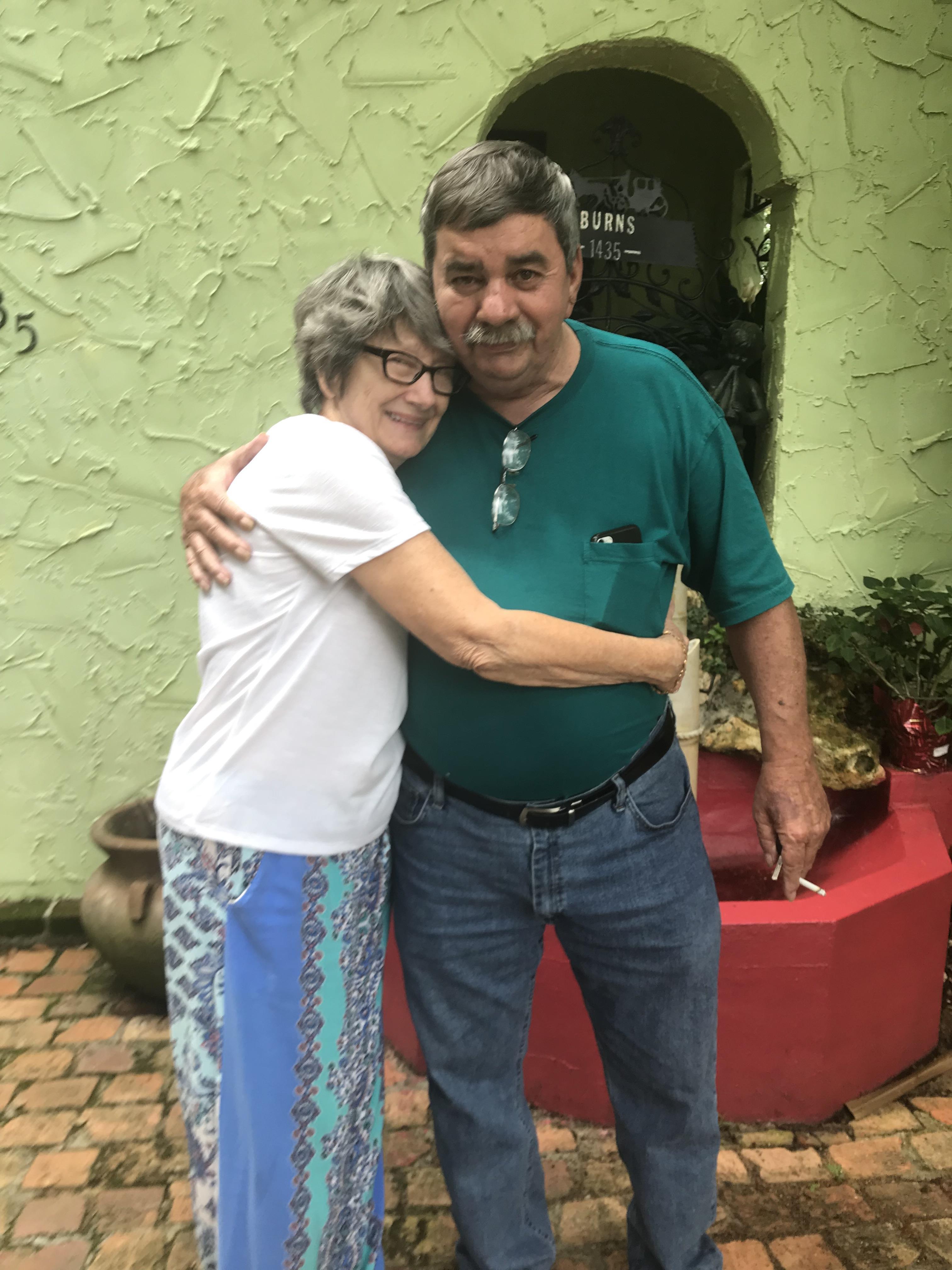 Two friends enjoy a heartfelt hug in a lively courtyard filled with greenery and colorful decor.
