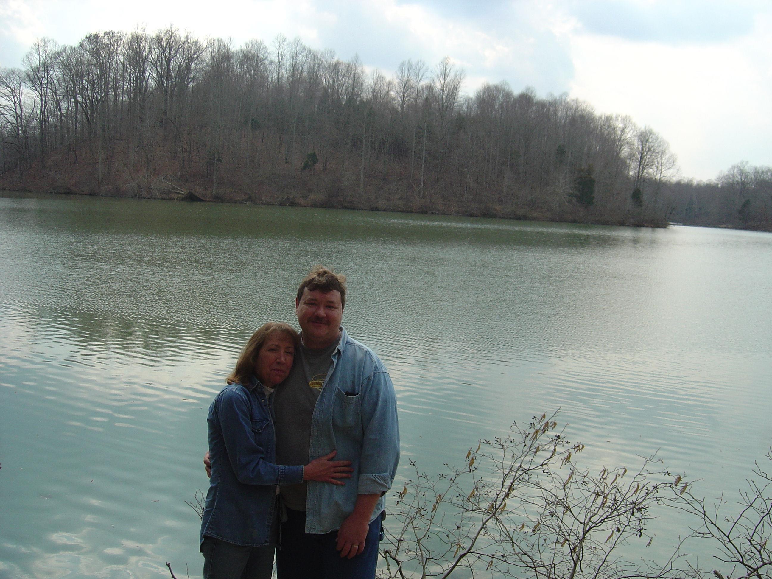 Two people embrace happily near a tranquil lake, with trees barely sprouting new leaves.