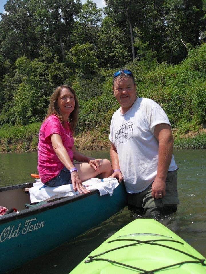 A couple relaxes in a canoe on a river, enjoying a sunny day amid beautiful natural scenery.