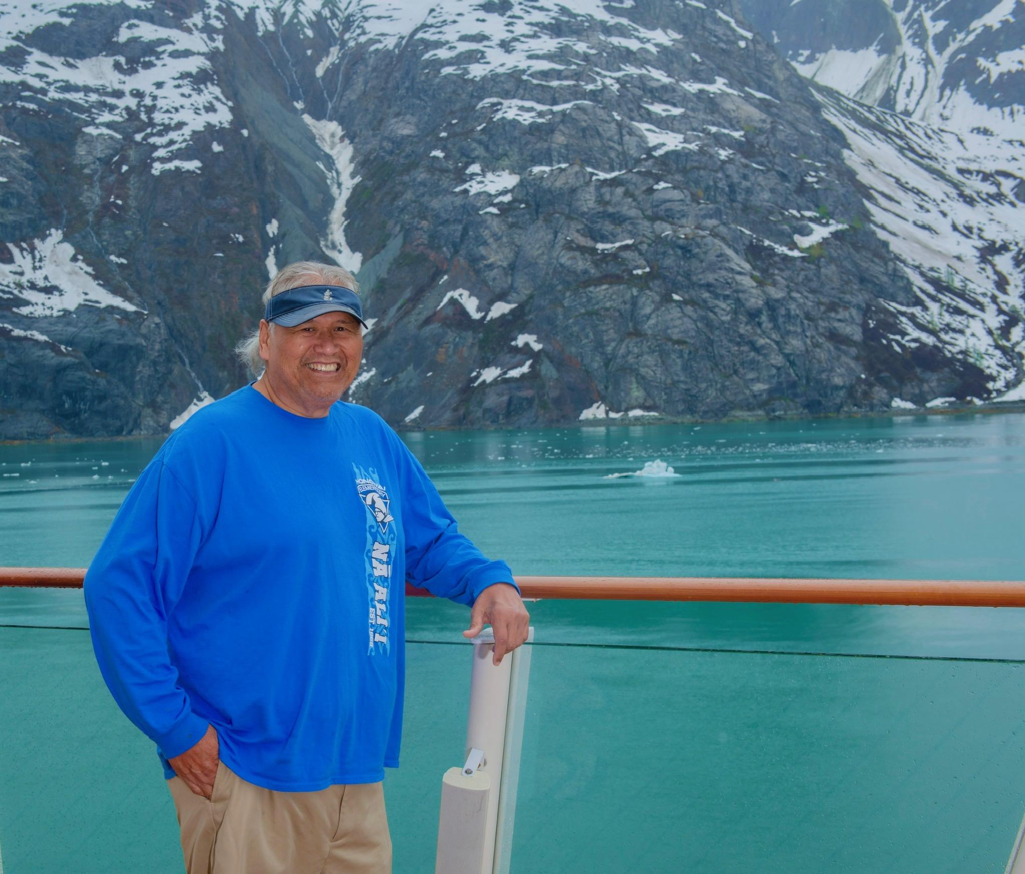 A man smiles on a cruise ship deck, admiring the calm glacial waters and mountains.