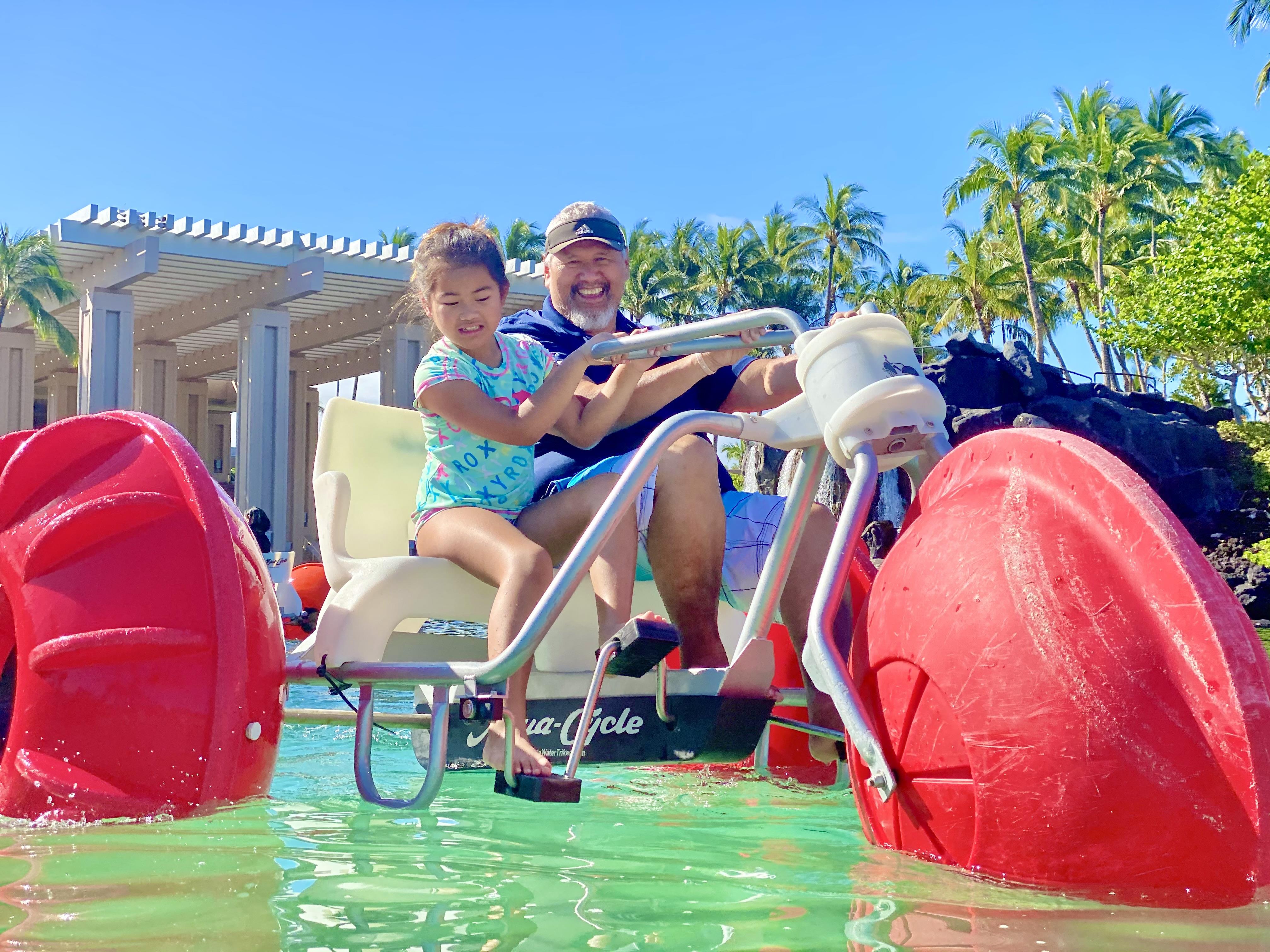A child and adult pedal together on a watercraft, surrounded by green water and palm trees.