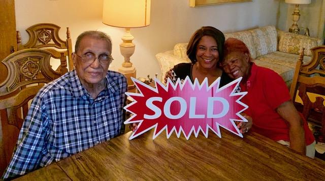 Three family members joyfully hold a sold sign in a warm and inviting living room setting.