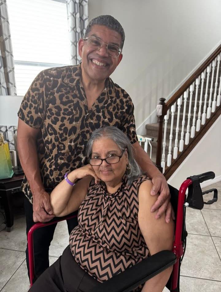 A man stands next to a woman in a wheelchair, both smiling warmly in a home interior.