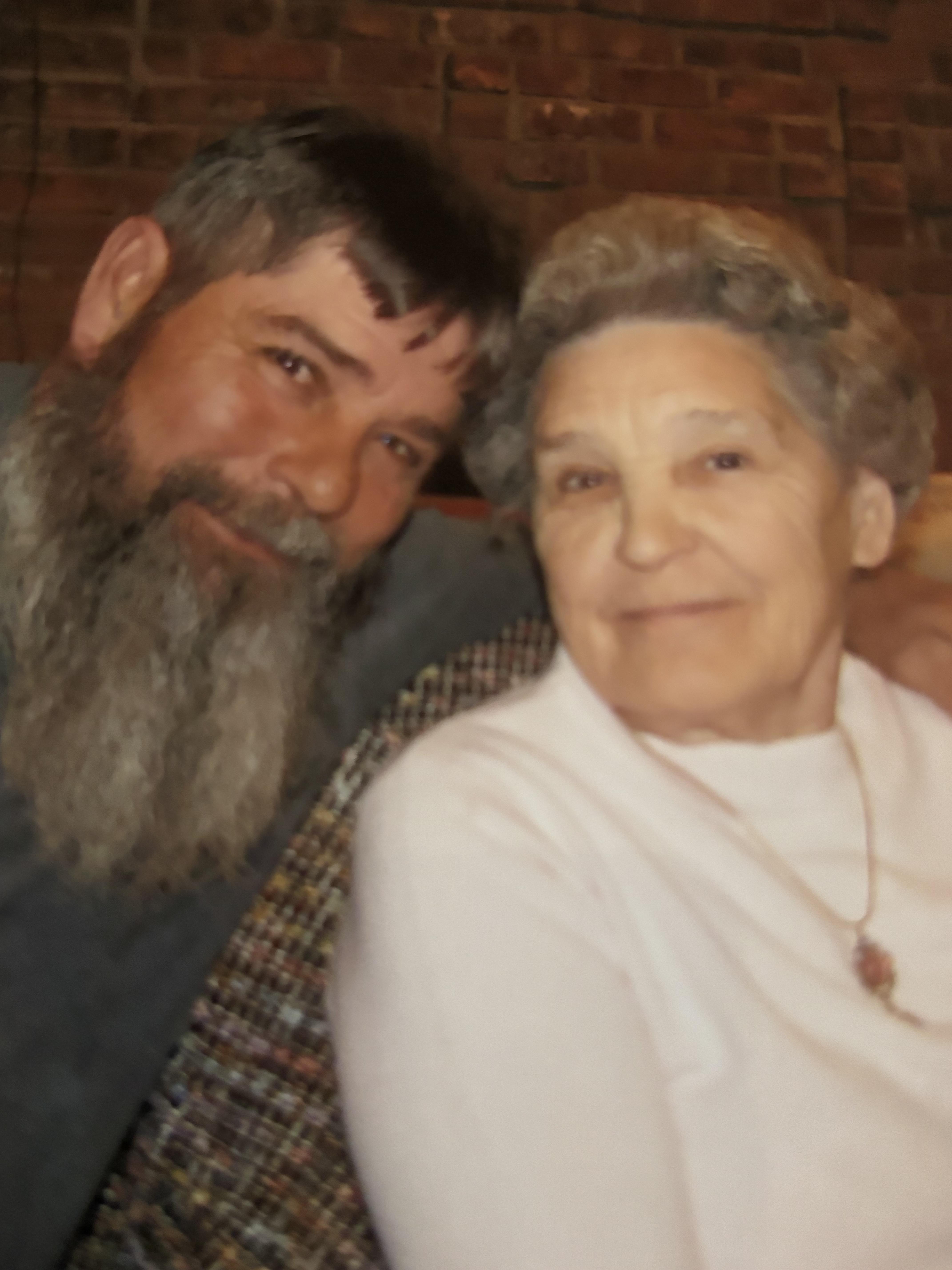A bearded man and an older woman share a joyful moment indoors, smiling at the camera.