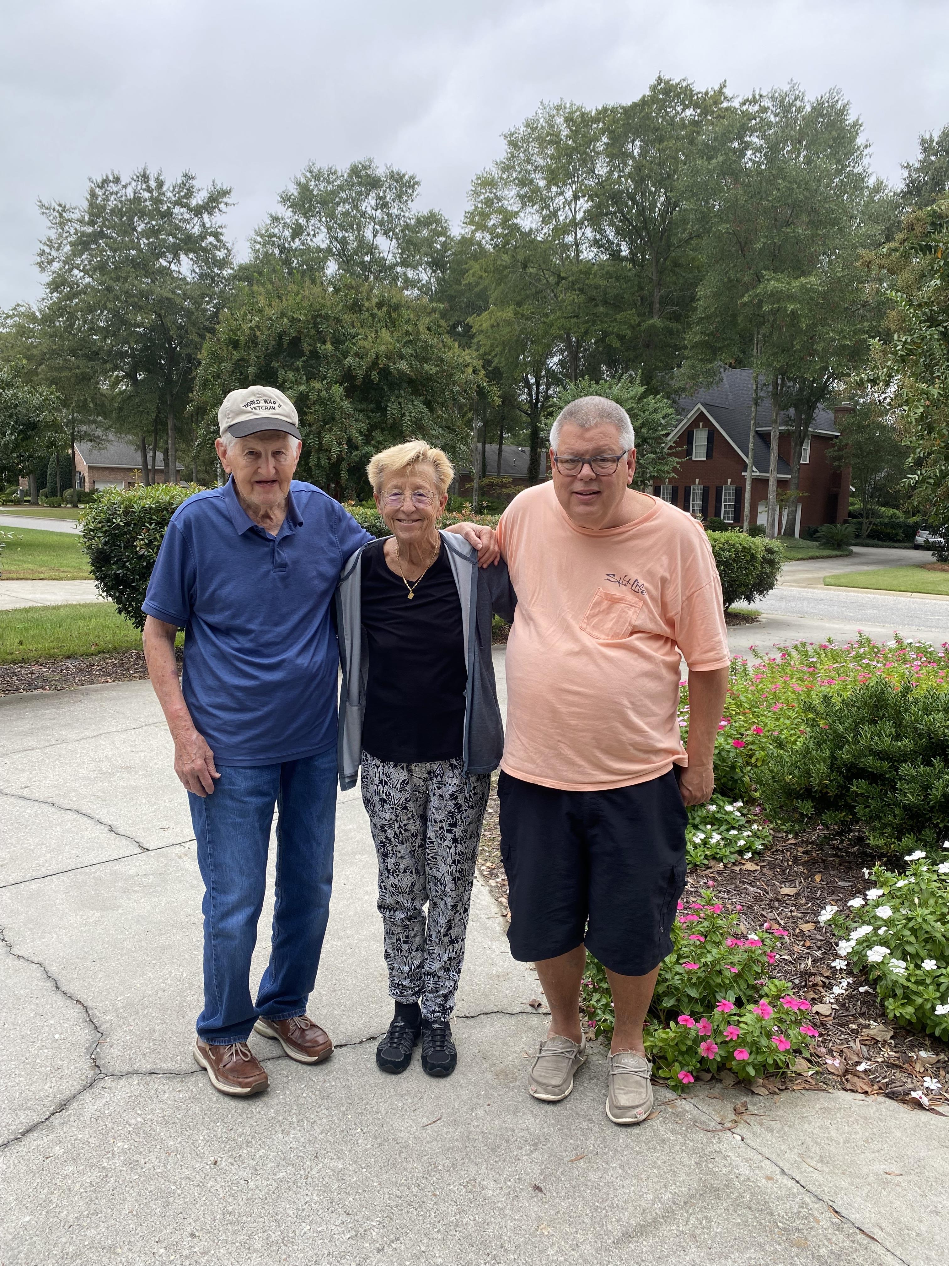 Three seniors smile while standing together in front of a house surrounded by greenery.