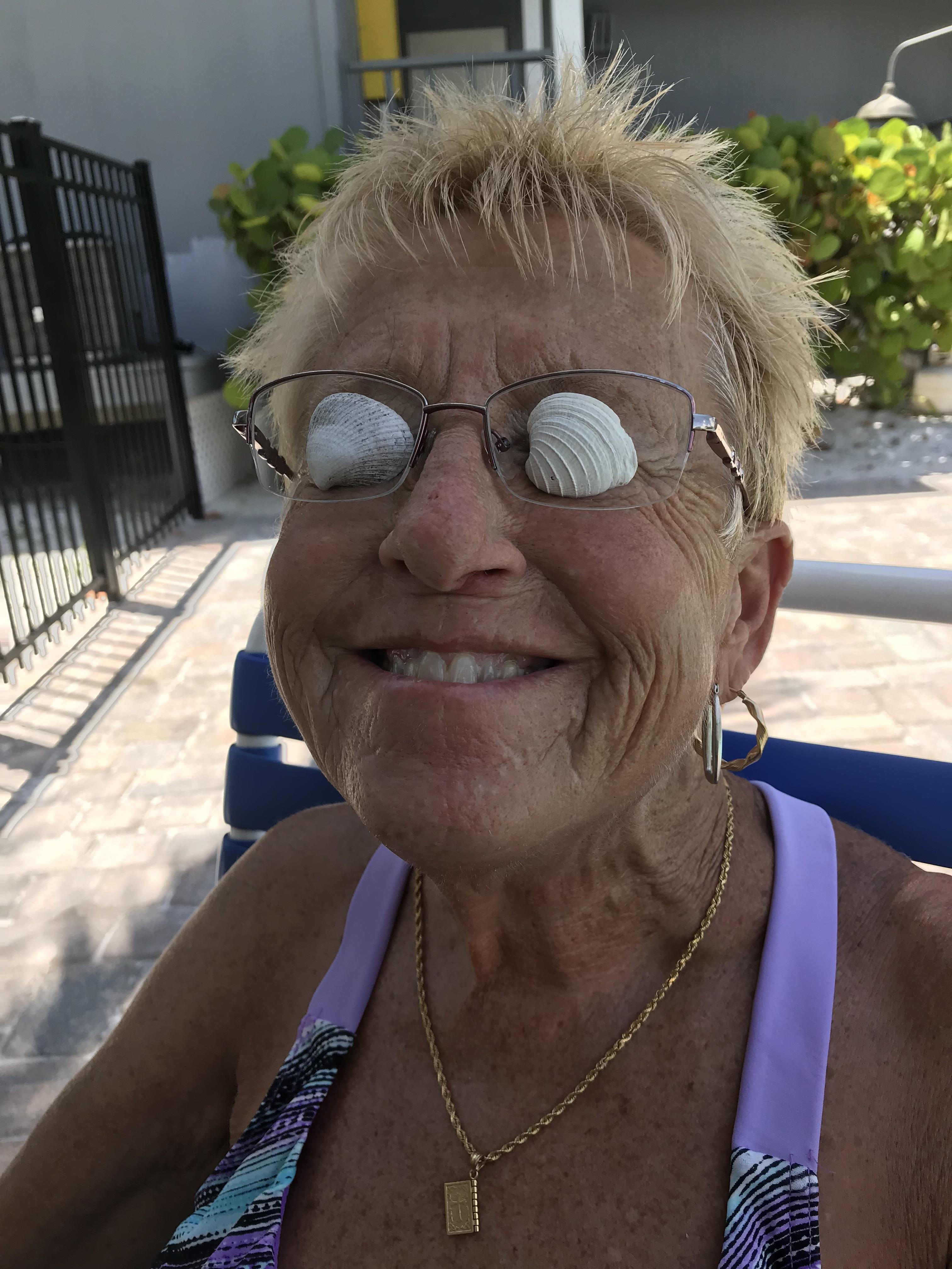 Cheerful elderly woman relaxes by the pool, wearing seashells as makeshift sunglasses under the sun.