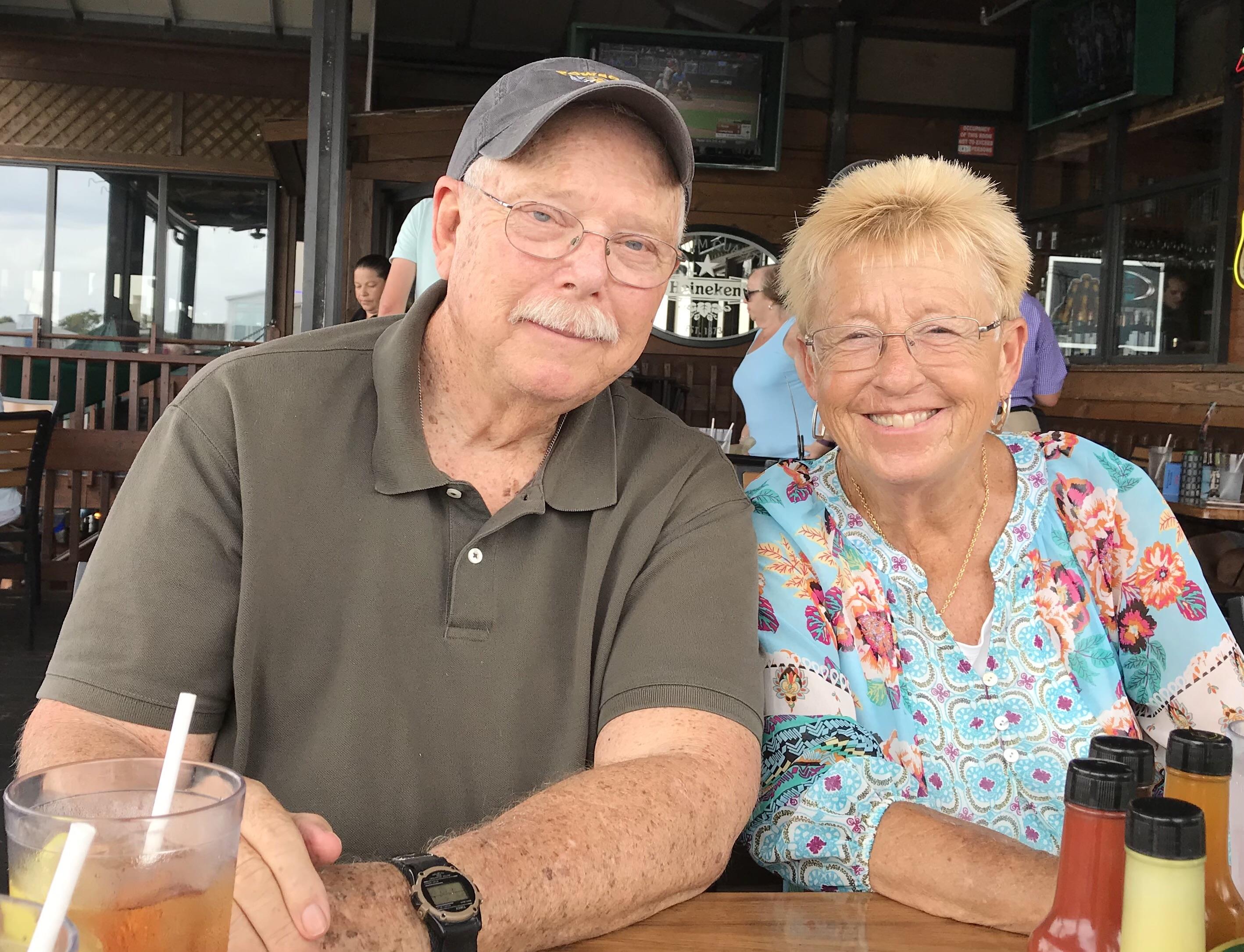 Elderly couple smiles over drinks at a vibrant waterfront restaurant with a relaxed vibe.