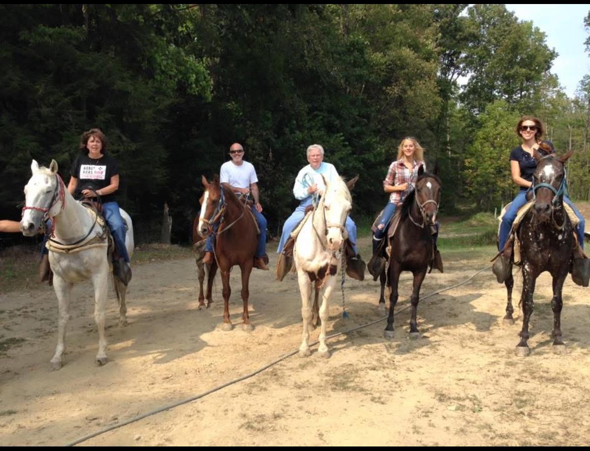 Five riders enjoy a horseback ride on a sandy path surrounded by trees in a rural setting.