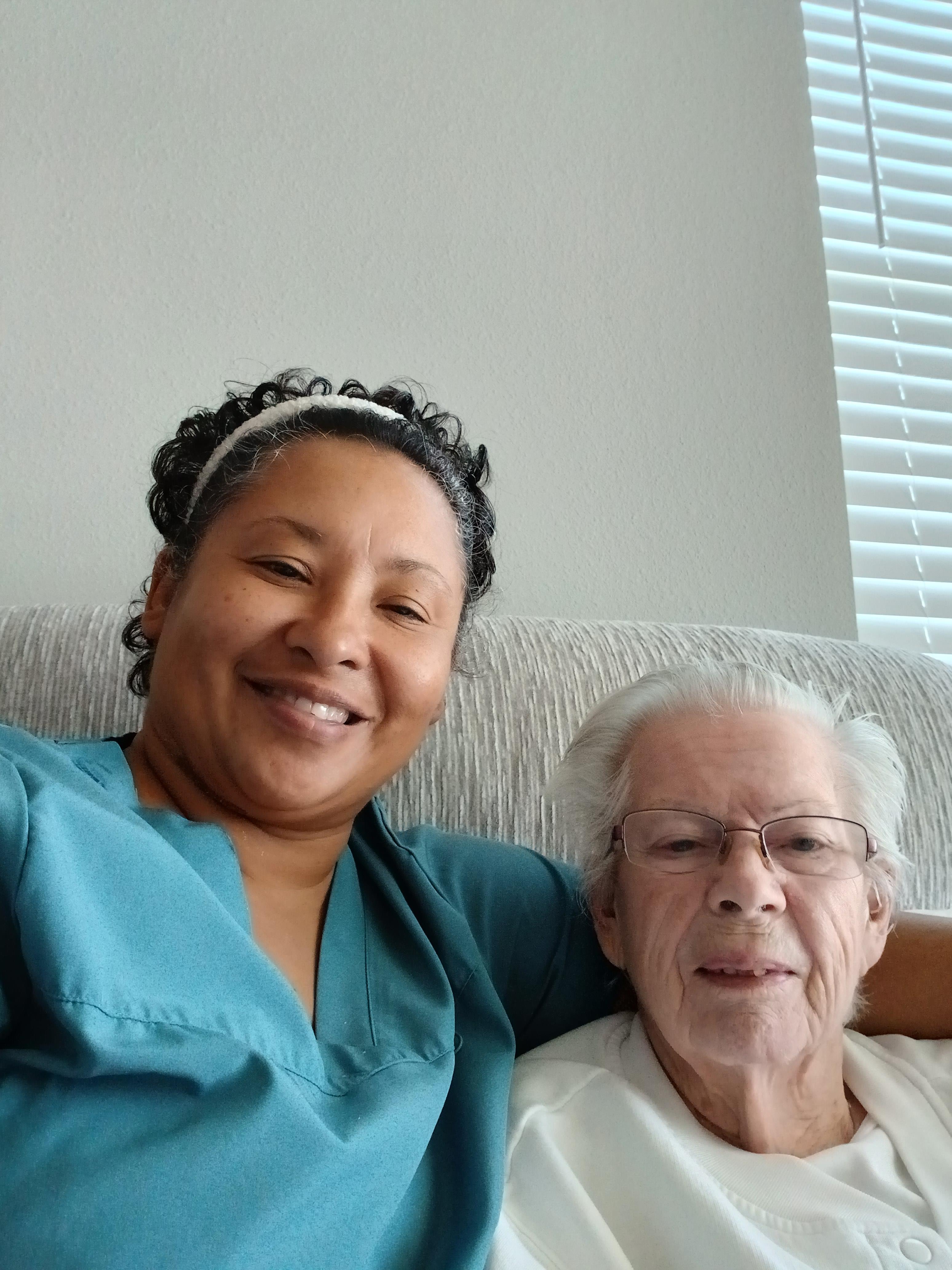 Caregiver and elderly woman smile together while relaxing on a couch in a bright living room.