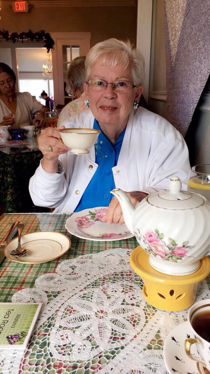 Elderly woman holds a teacup while sitting at a table with tea service and dessert.