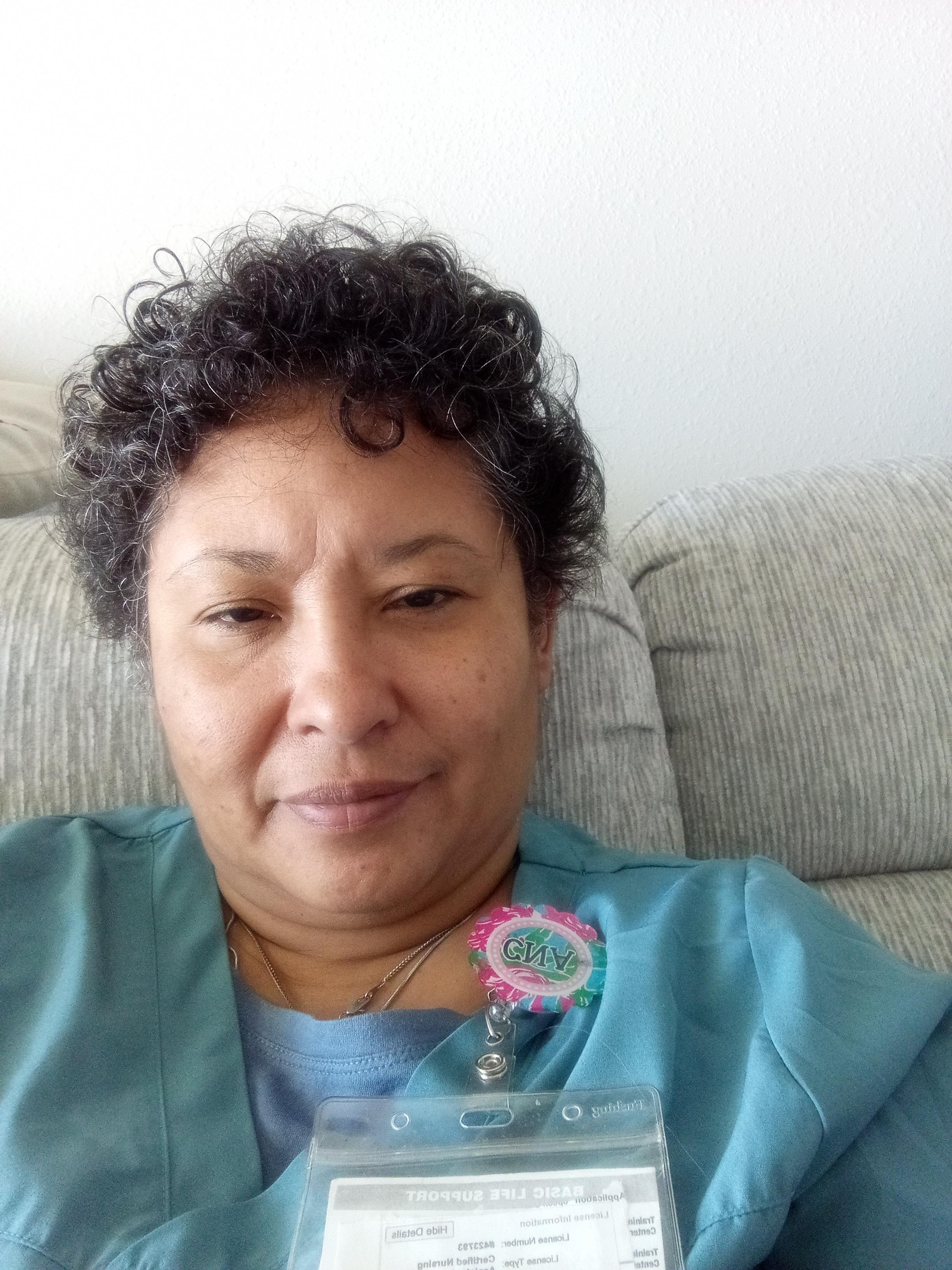 A woman with curly hair is sitting comfortably on a light-colored couch, smiling gently.