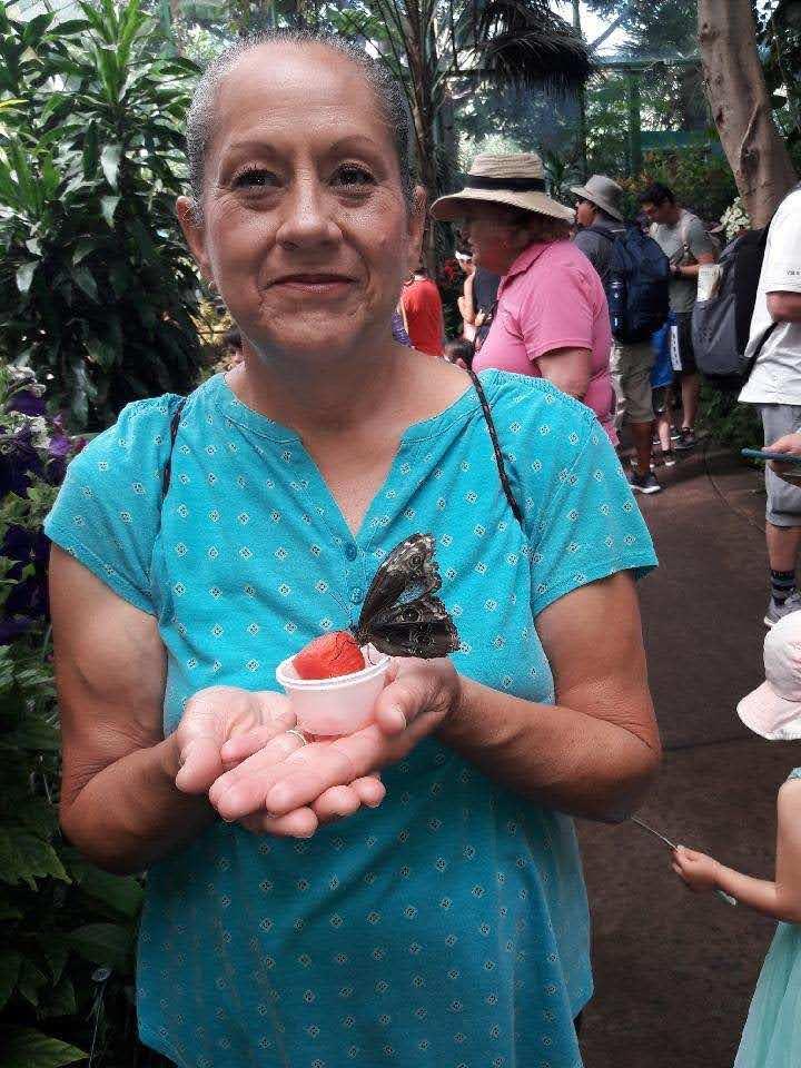 Woman smiles as she holds a butterfly on her hand near a group of visitors at a garden.