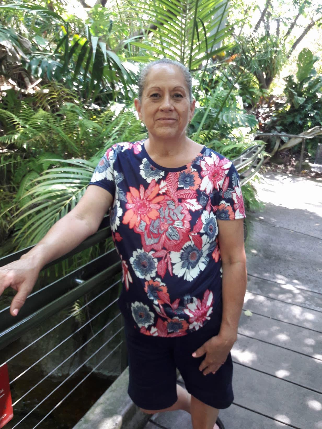 An elderly woman stands confidently by a railing in a lush garden filled with green foliage.