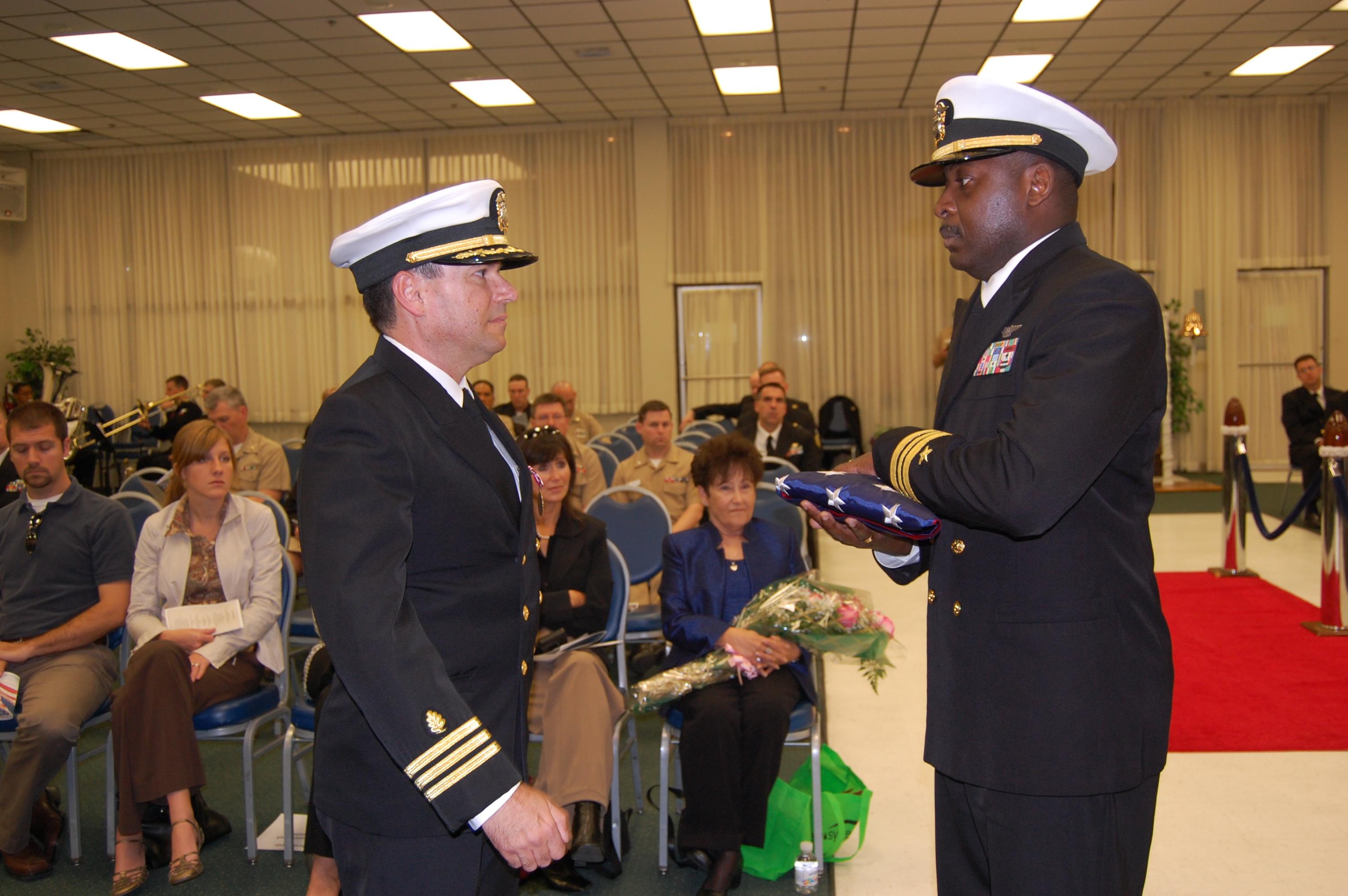 Two navy officers participate in a formal flag presentation ceremony while attendees observe.
