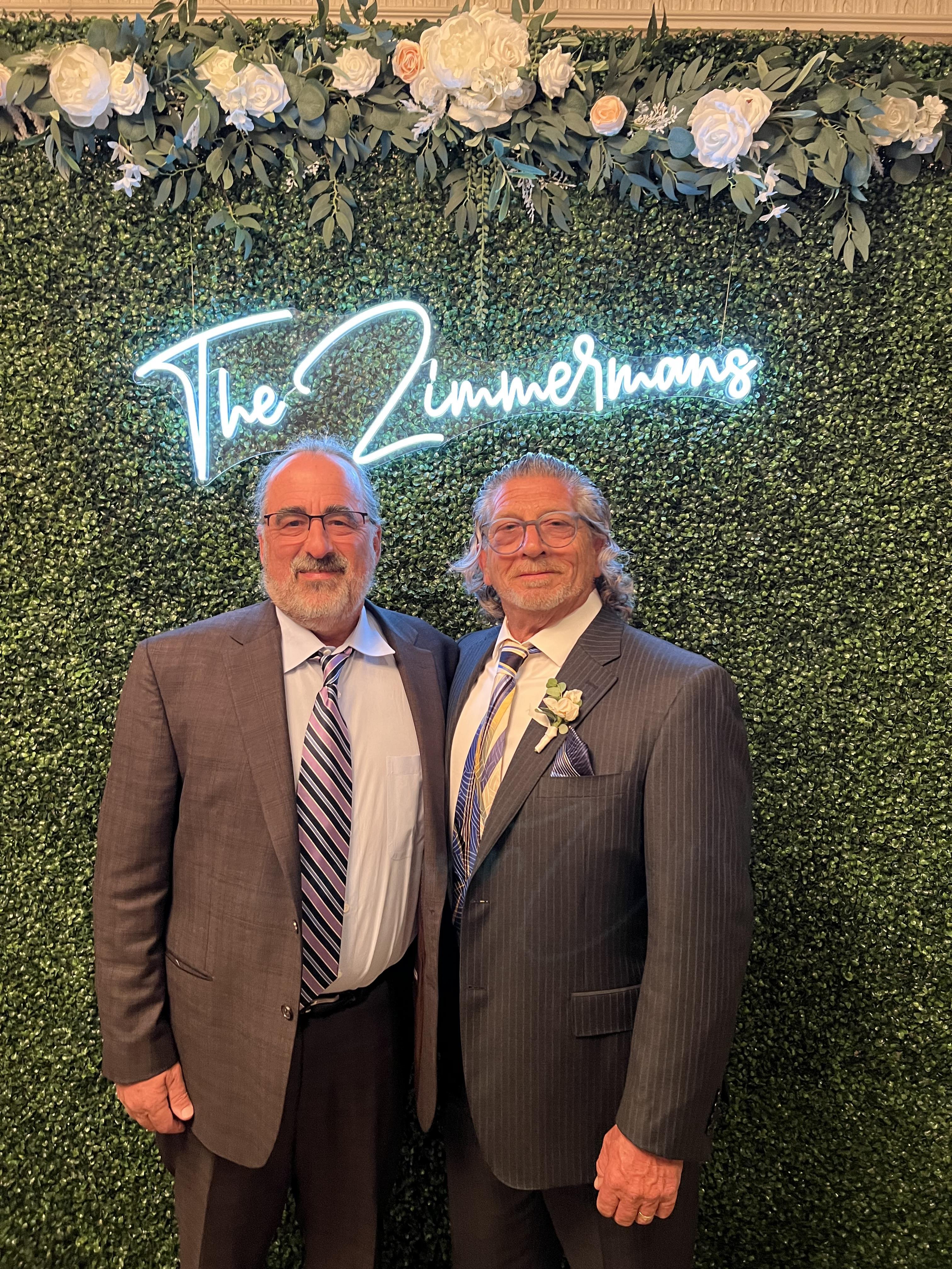 Two men in formal attire smile for a photo against a lush green backdrop during a gathering.