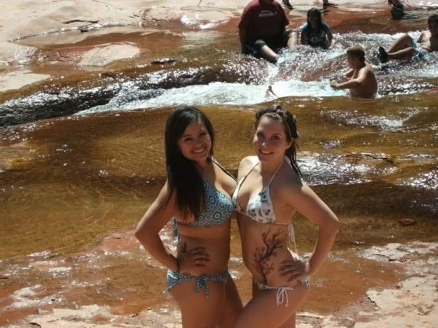 Two friends pose happily in bikinis while enjoying a day at a sunny swimming hole with others.