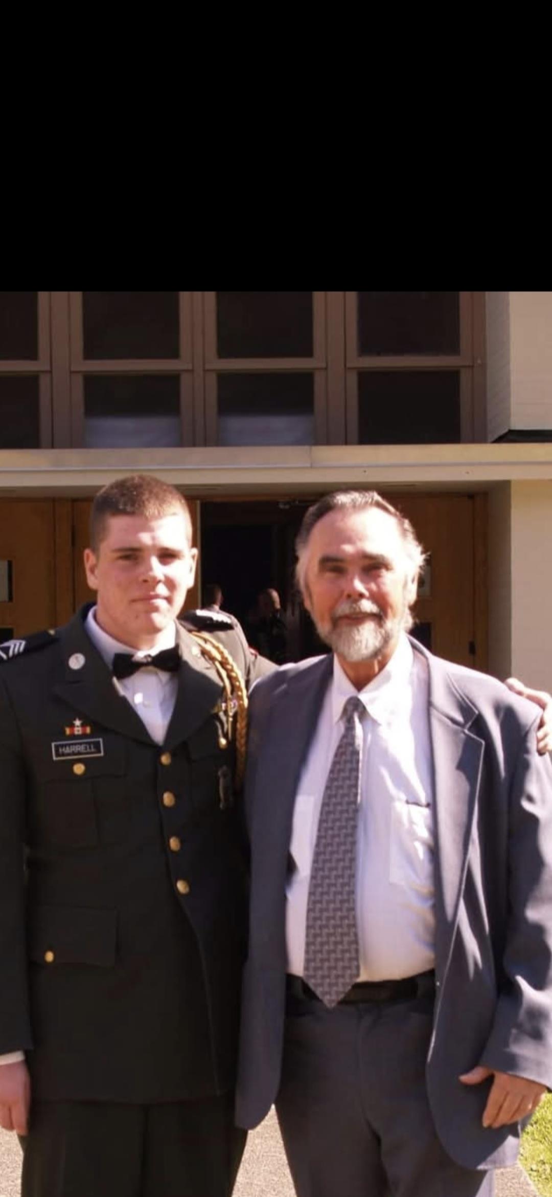 Two men stand side by side, smiling proudly in formal attire outside a distinguished building.