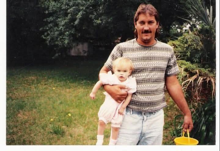 A man carries a young child in a garden setting, surrounded by greenery and sunlight.