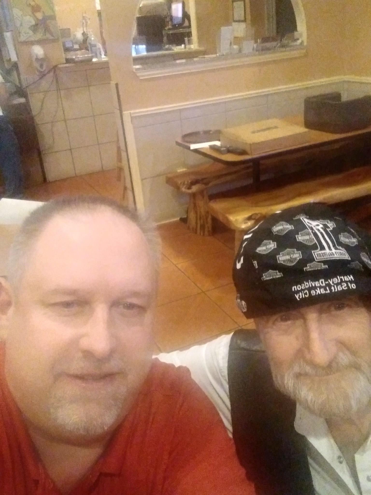 Two men smile as they take a selfie in a cozy restaurant filled with wooden furnishings.