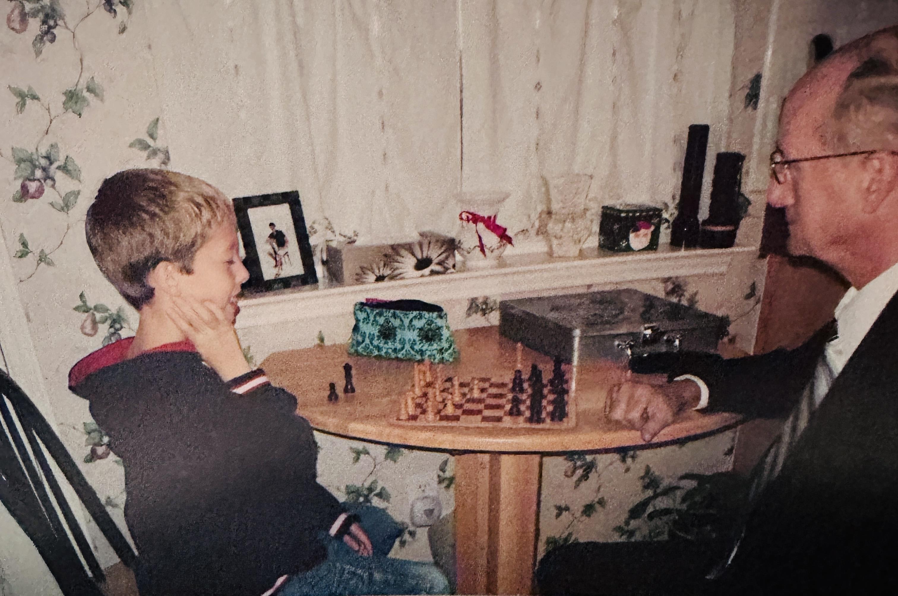 A young boy and an elder engaged in a chess match at a wooden table in a warm, inviting living room.