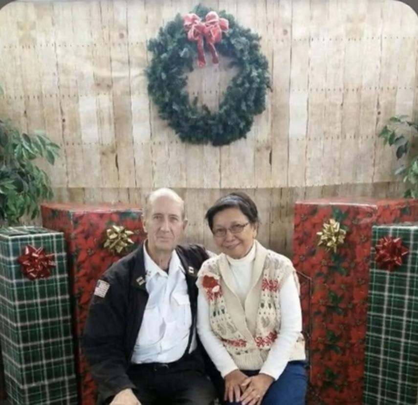 A couple sits together near decorated Christmas gifts and a festive wreath in a cozy setting.