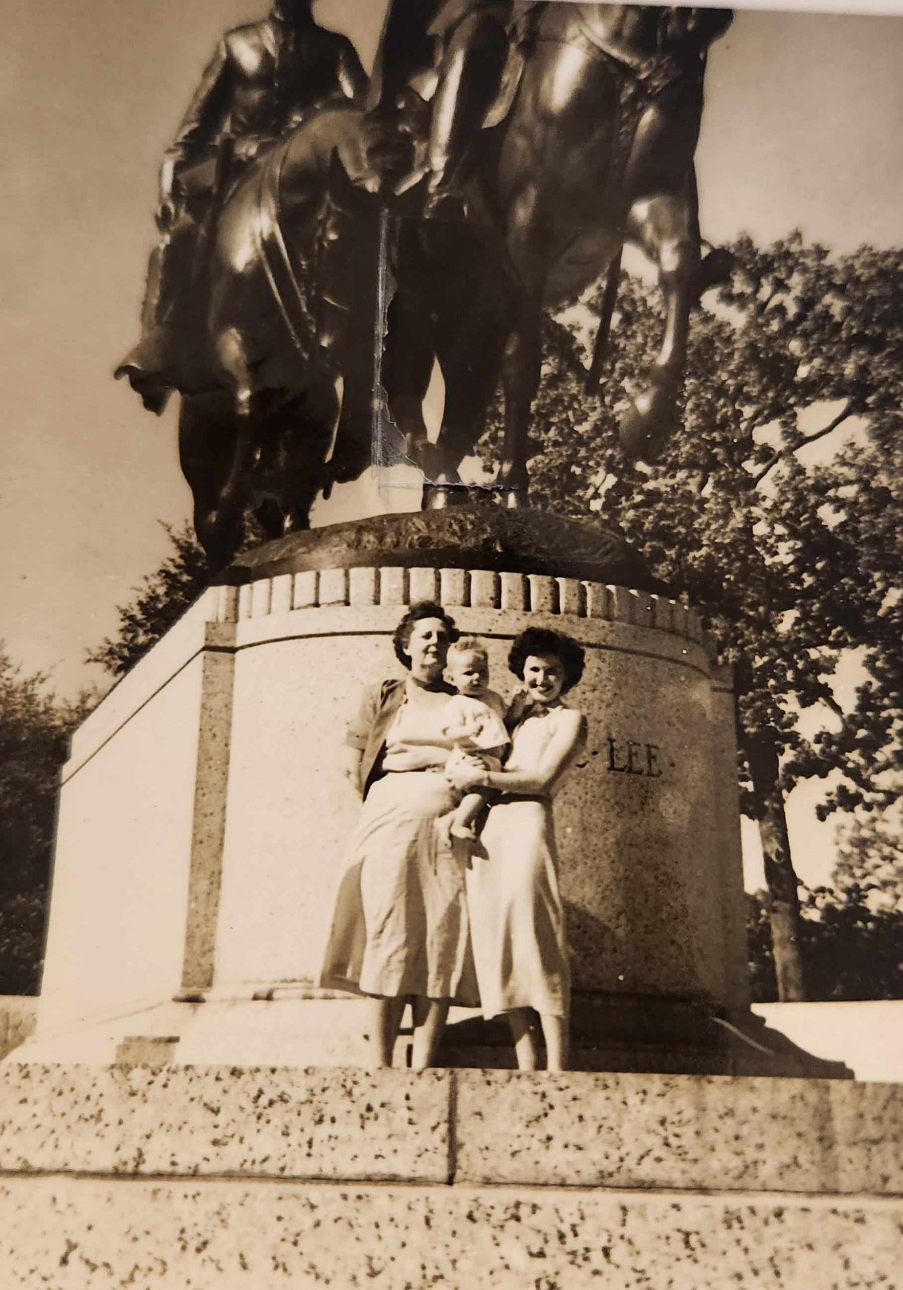 Two women and a child stand near a large statue in a public park, enjoying a sunny day together.