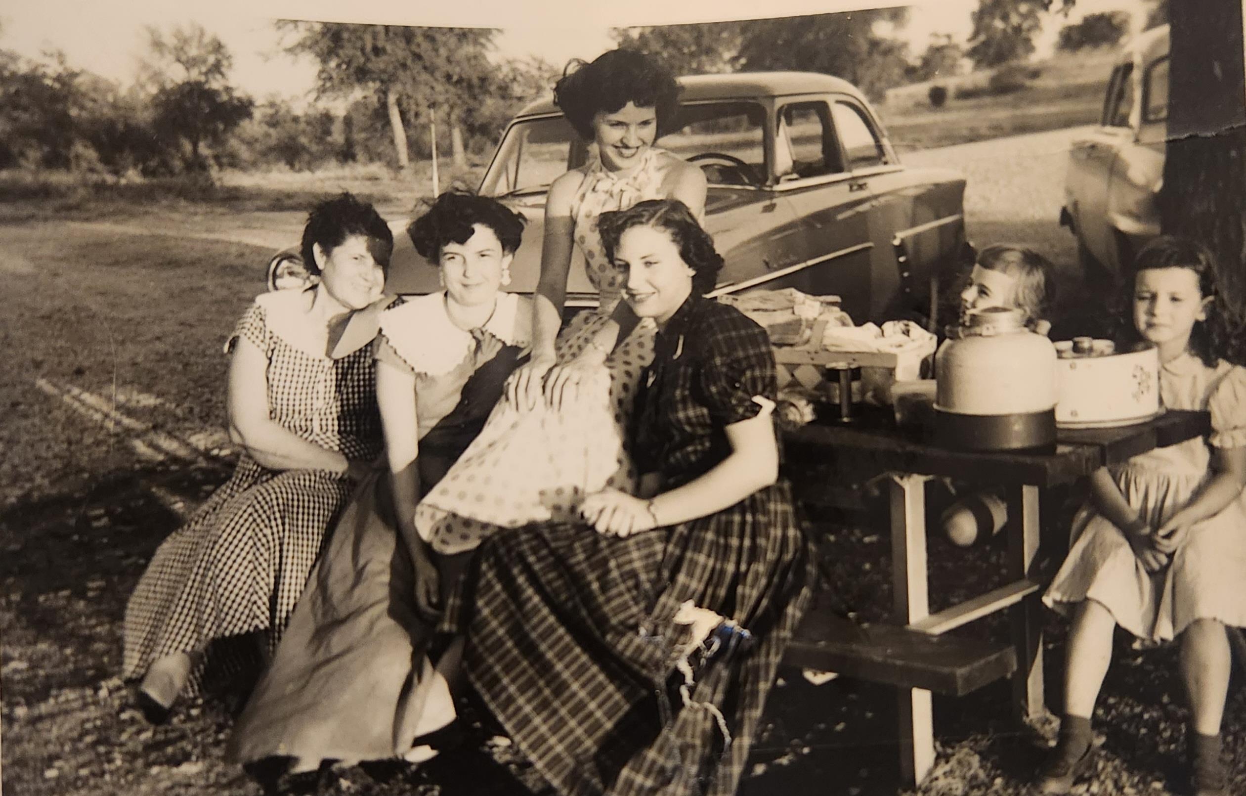 Four women gather around a picnic setup, smiling and enjoying the sunshine by a classic car.