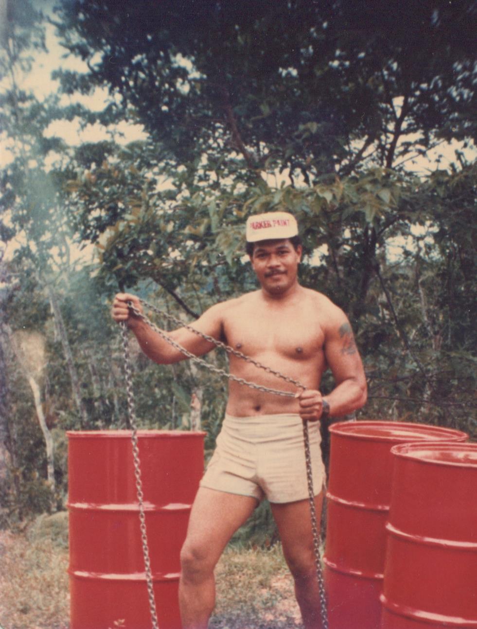 A shirtless man with a strong build holds chains near red barrels surrounded by trees.