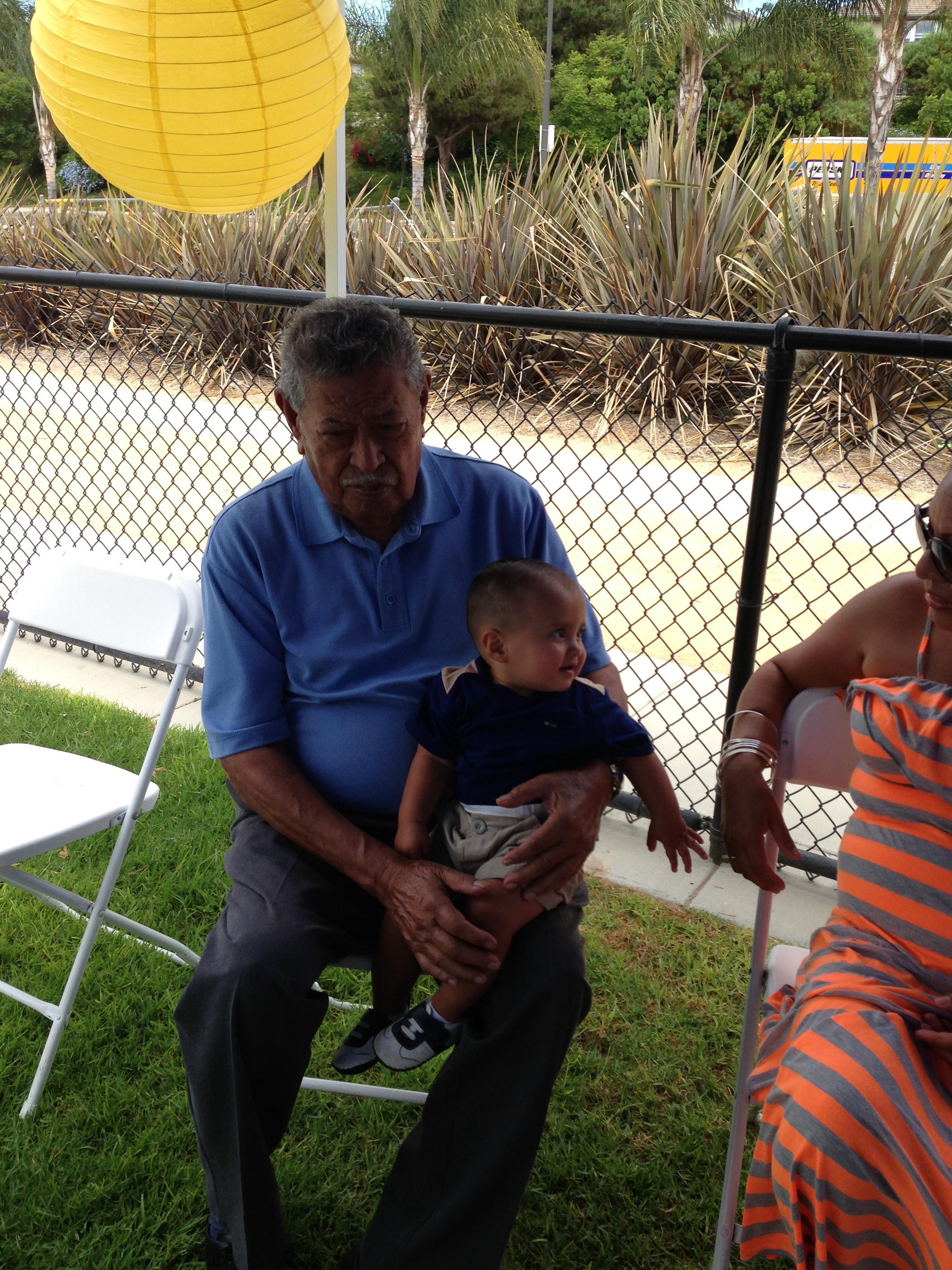 An elderly man sits with a young child in his lap during a warm family gathering in daylight.