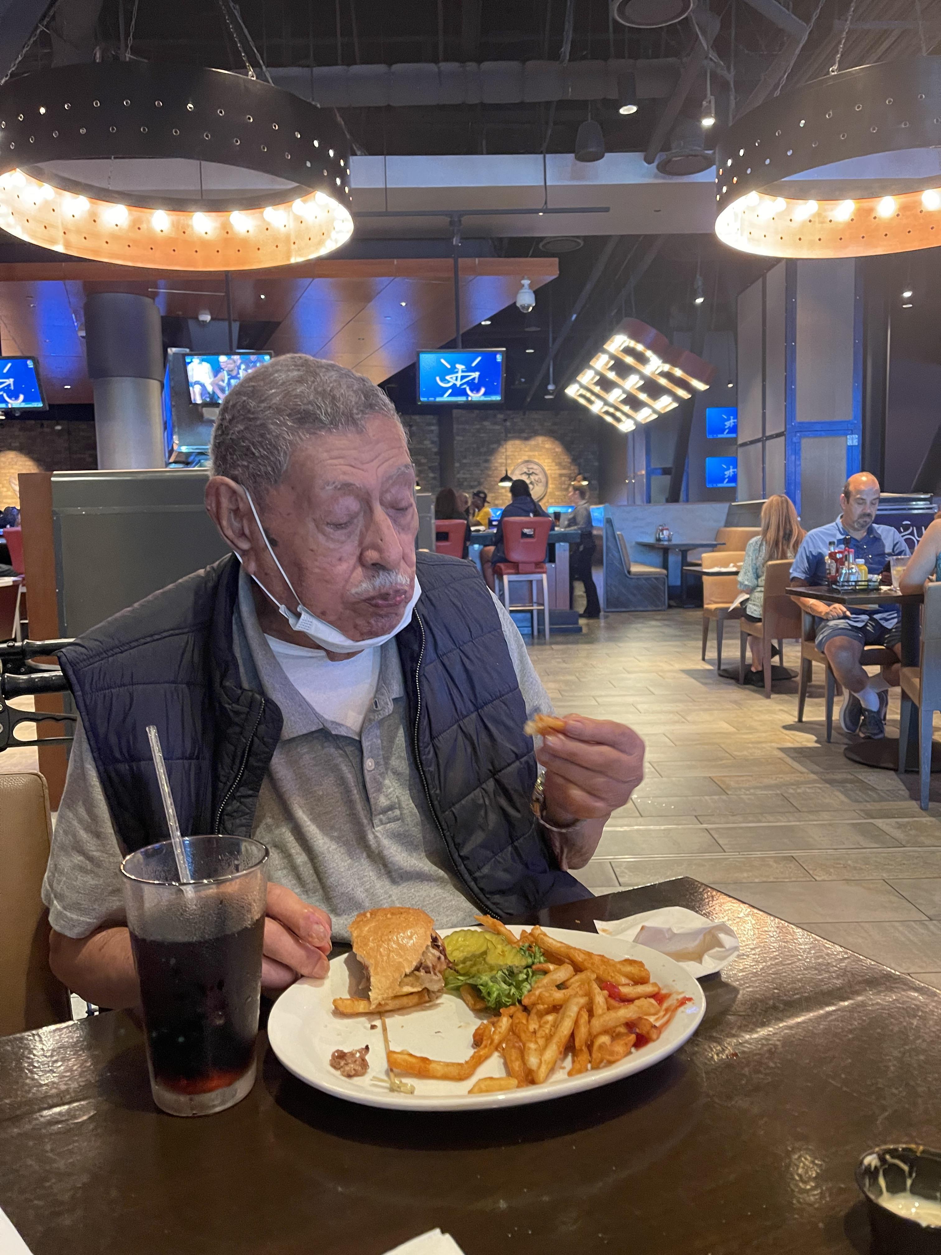 An elderly man sits at a table savoring a burger and fries while surrounded by diners.