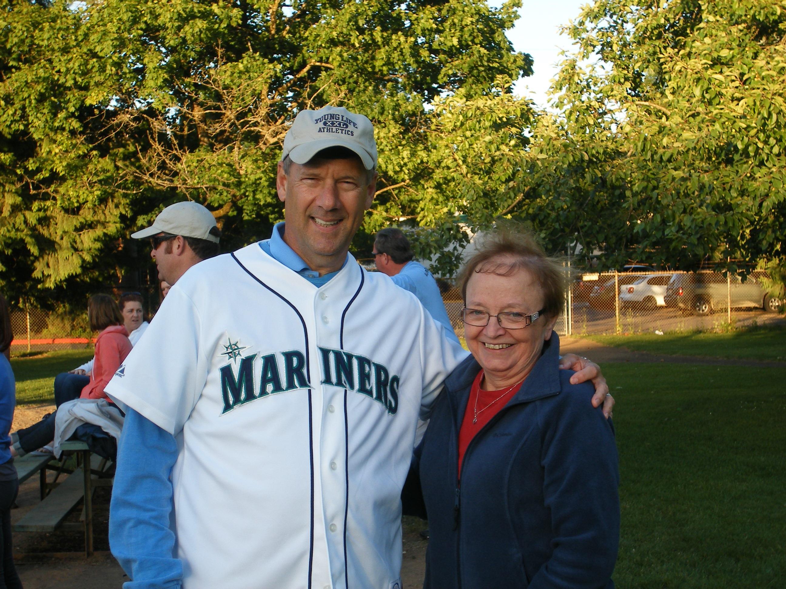 A man wearing a baseball jersey poses with a woman in a park at dusk, both smiling happily.