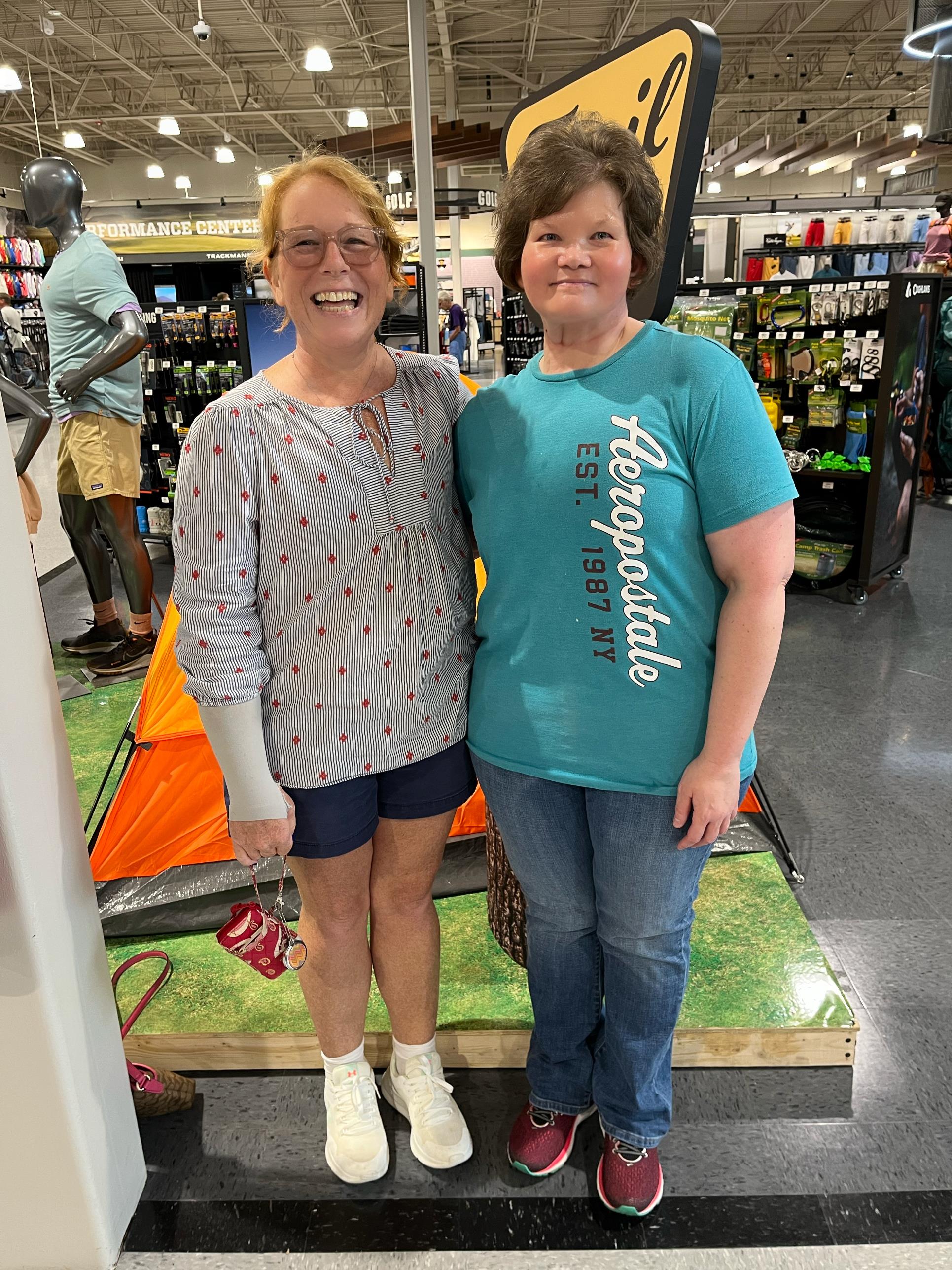 Two women smile happily next to a colorful camping tent in a retail store.