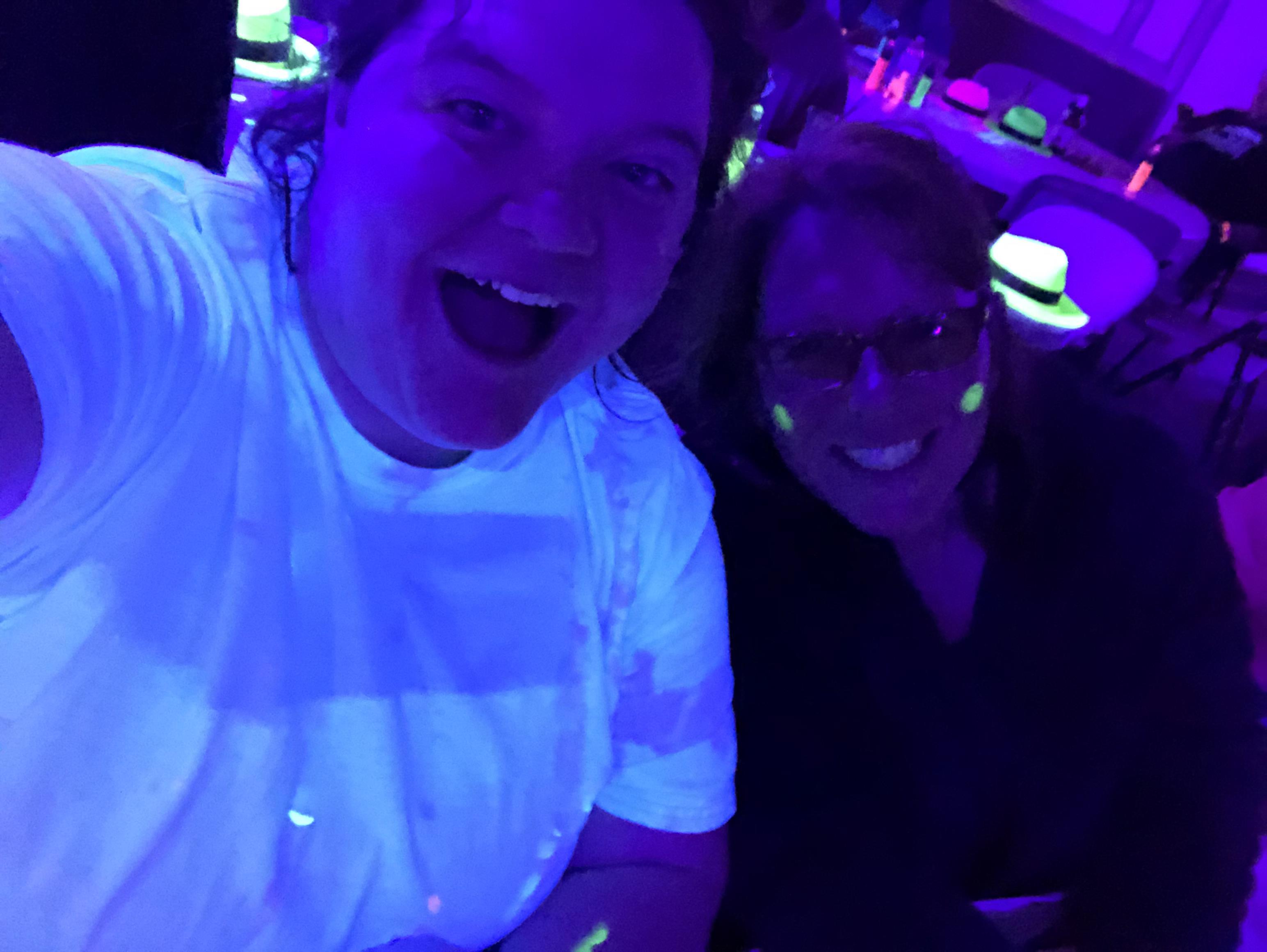 Friends smile and pose for a selfie amid colorful neon lighting at a lively club.
