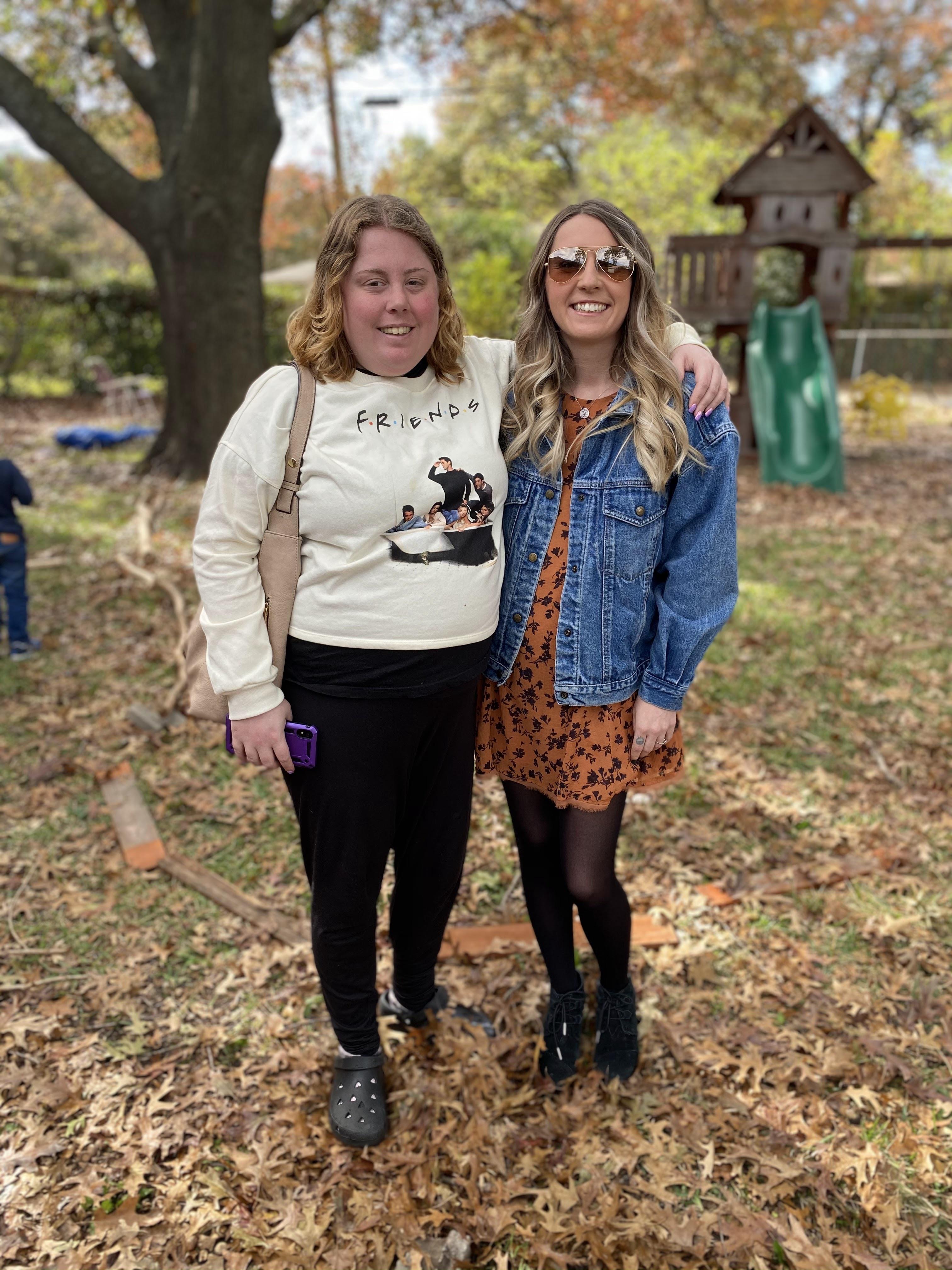 Two women pose happily in a park surrounded by colorful fall leaves and playful children.