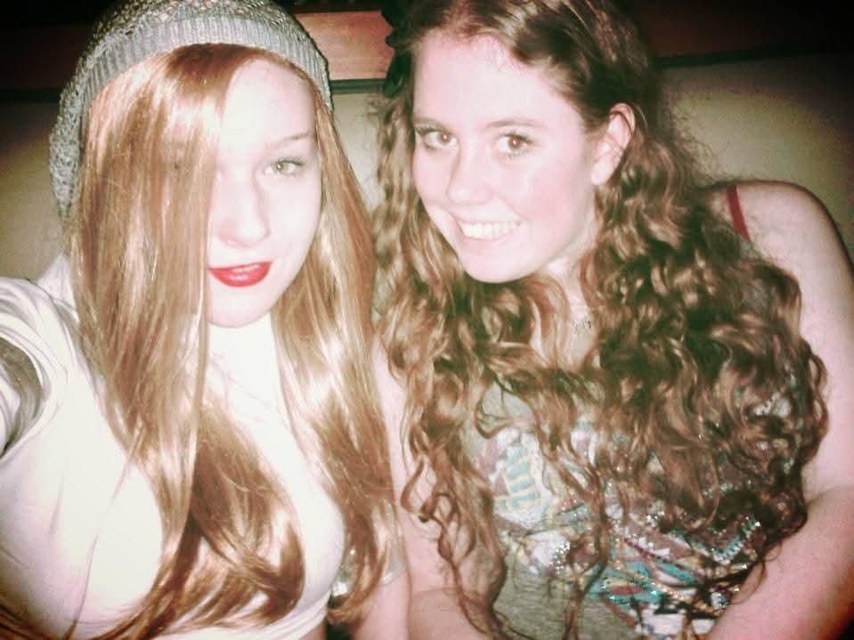 Two friends with long hair are smiling, dressed casually, enjoying their time together at a bar.