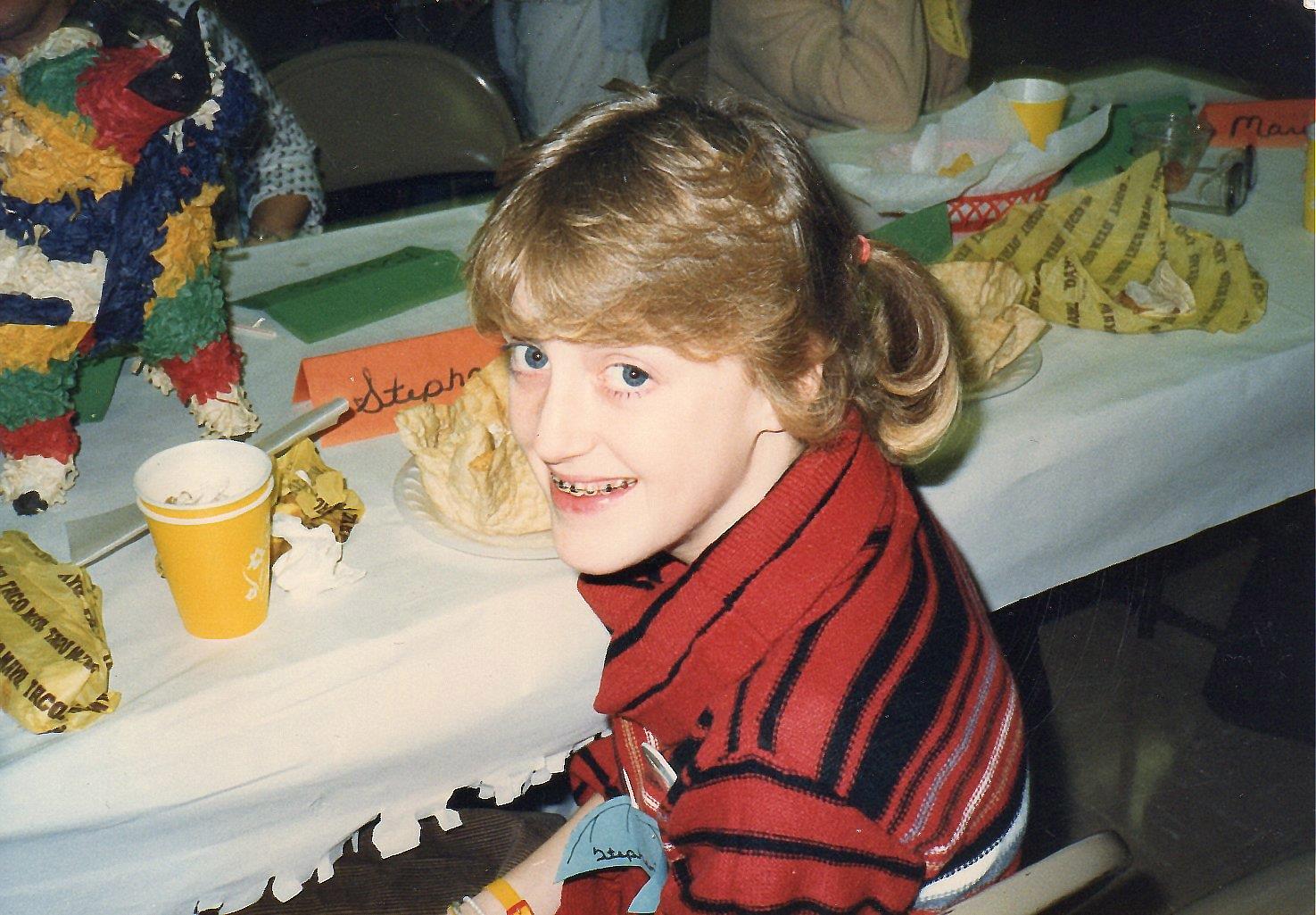 A young girl smiles while sitting at a table filled with colorful decorations and food, having fun.