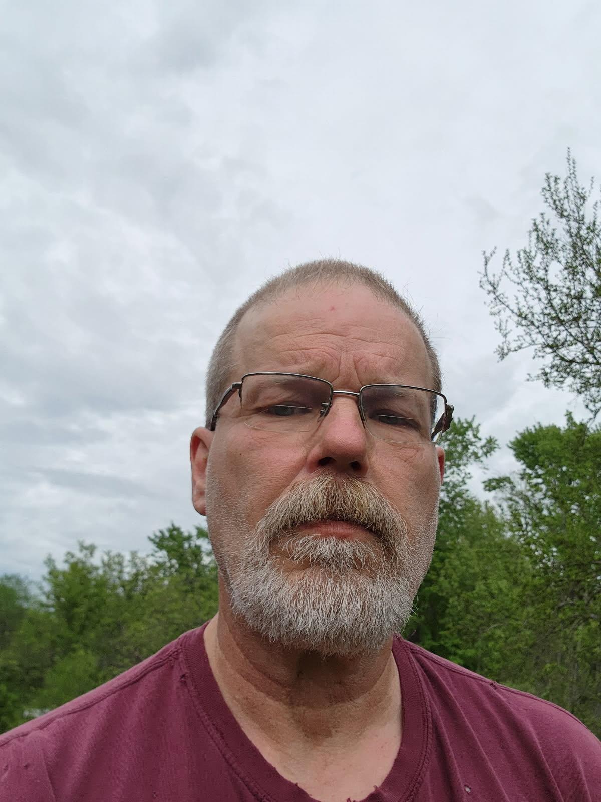 Man with a beard and glasses poses in a green environment on an overcast day.