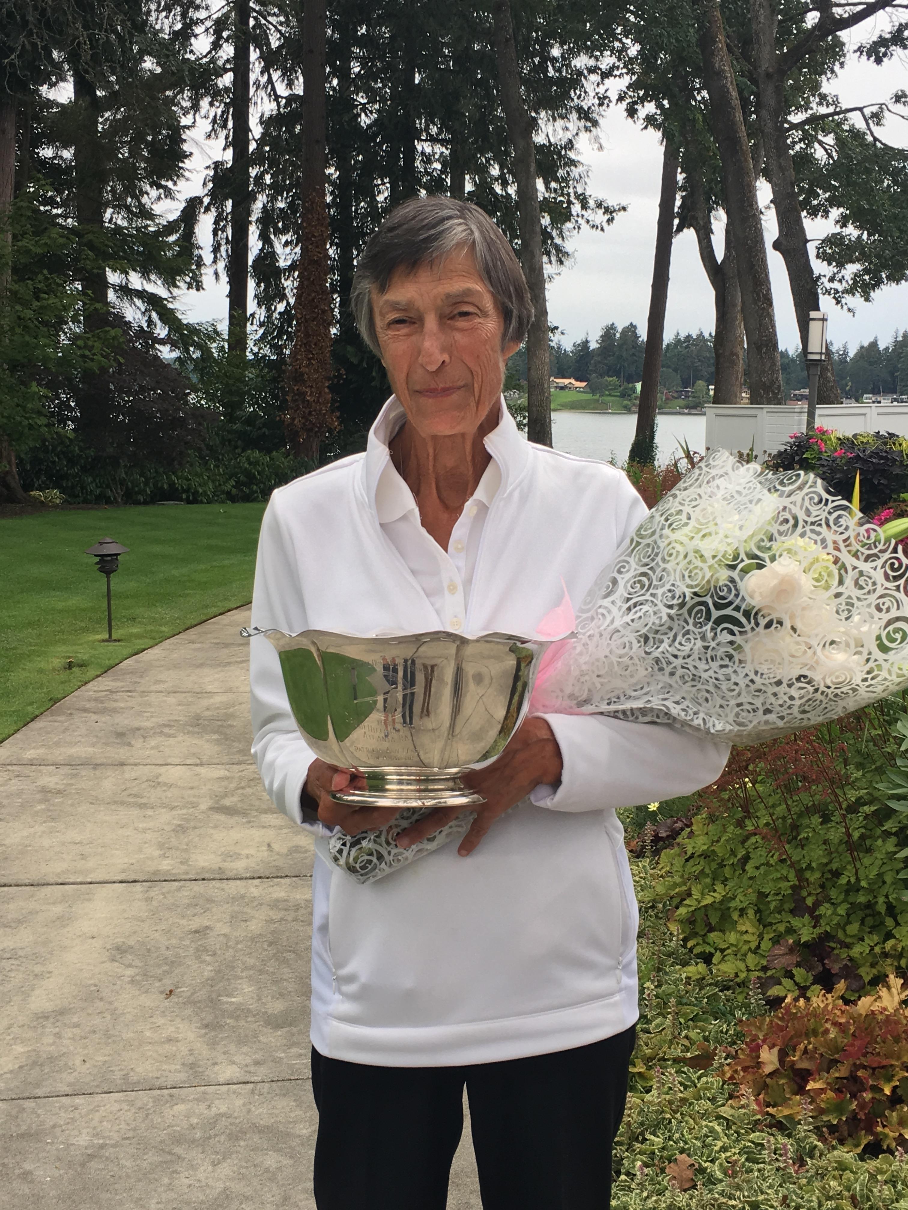 Person stands proudly with a trophy and bouquet near a serene lake surrounded by greenery.