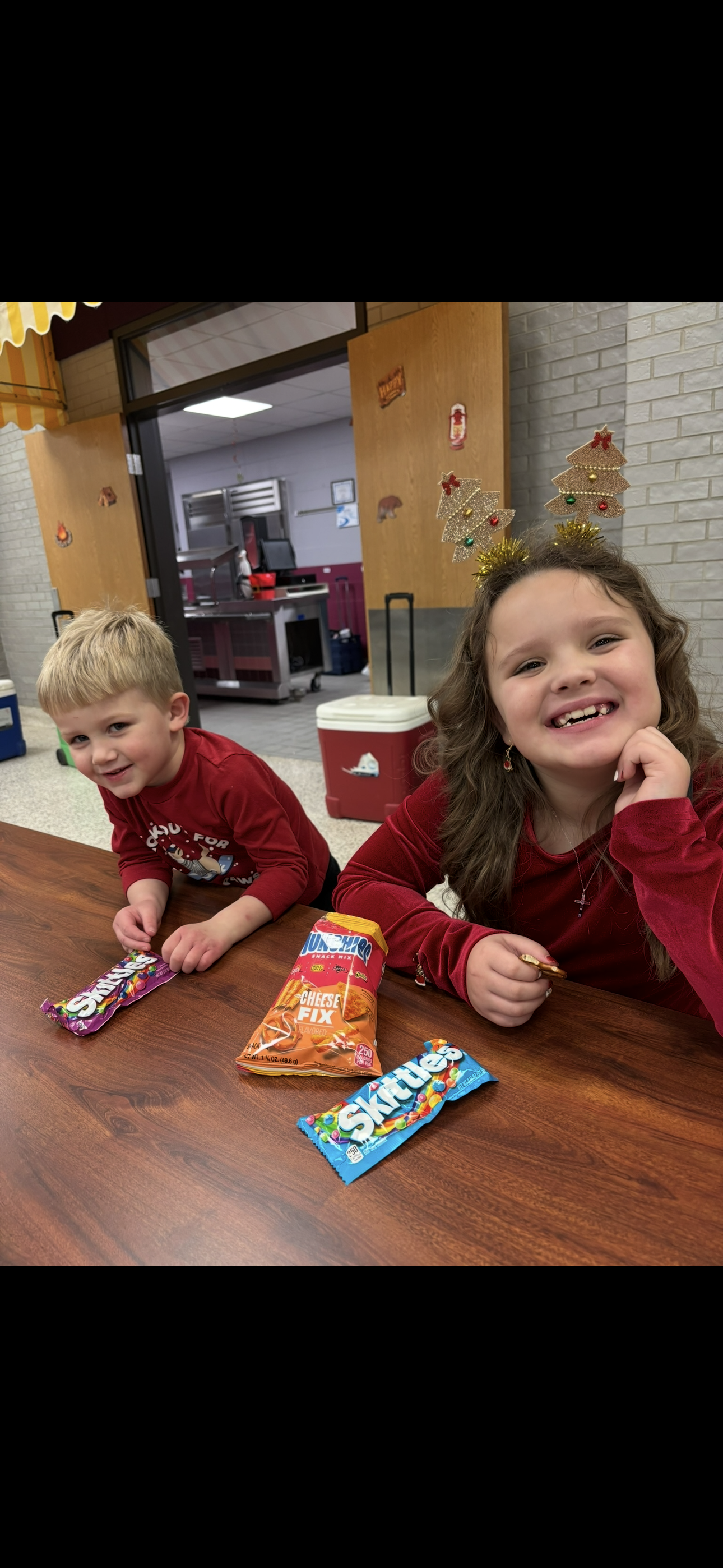 Two children sitting at a table, smiling as they eat snacks during a holiday celebration.
